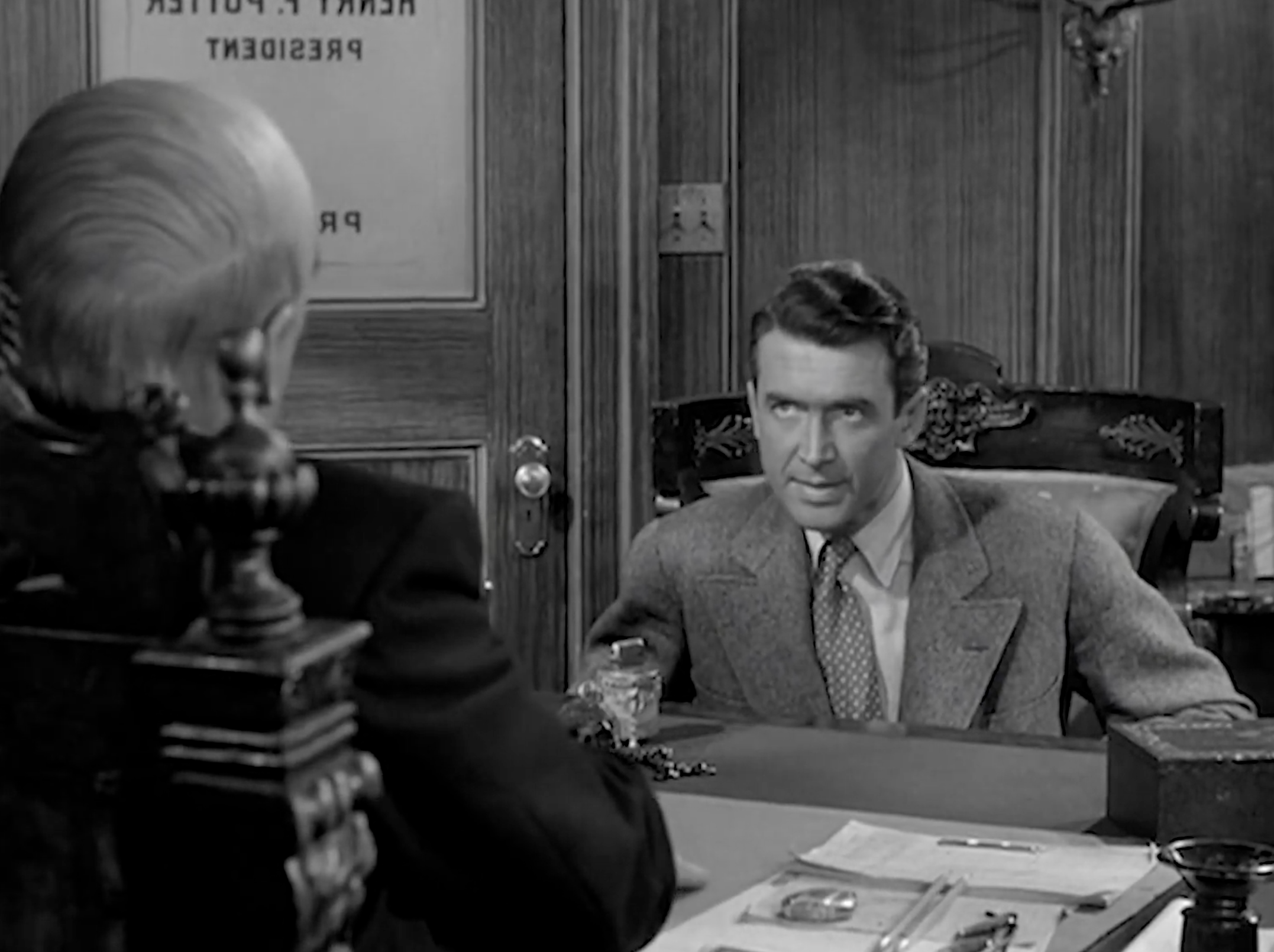 Weakness on Display This reverse angle puts George where Potter sat before—centered, framed by the heavy chair and the office door—yet it doesn’t play as a power shot. His body leans forward, eyes locked, caught between curiosity and conviction. With Potter out of focus in the foreground, the camera exposes George’s inner struggle: this is what temptation looks like from the outside. He’s not evil, just human—and the scene reminds us that moral strength isn’t the absence of weakness, but the choice to push through it.