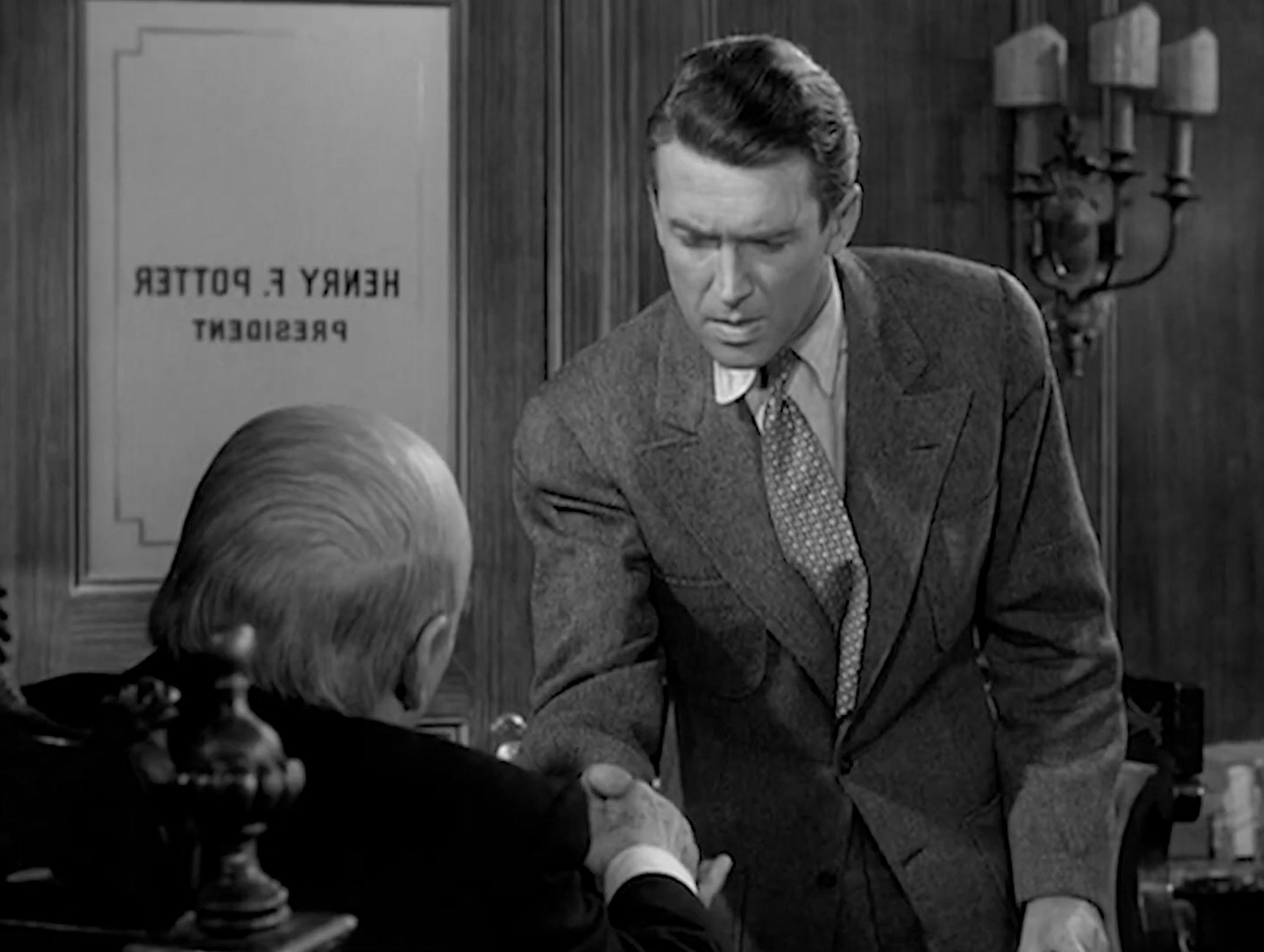 The Moment of Clarity The framing traps George between Potter’s hand and the office door that literally bears Potter’s name, as if he’s one handshake away from walking through it forever. The camera lingers on George’s downward glance, letting us see the thought process: this isn’t just a business agreement, it’s an alignment of character. By holding on this close, slightly high angle, the film turns a simple gesture into a moral pivot point—George sees the hand, recognizes the cost, and decides he can’t sell his soul for a salary.