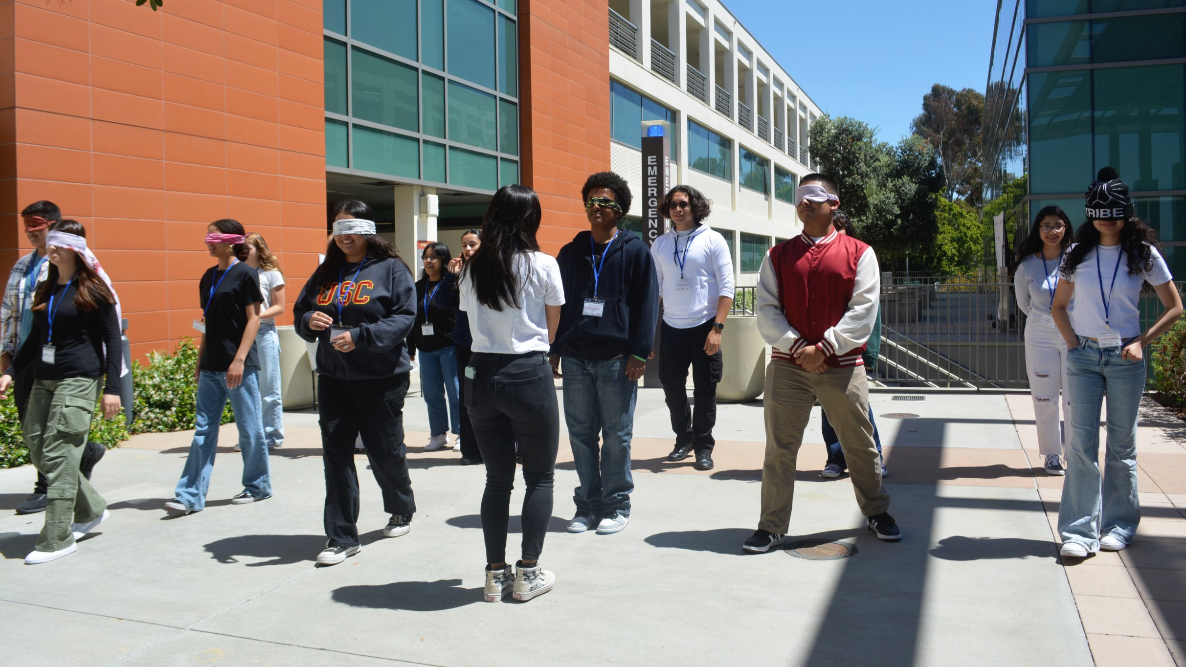 Youth participate in an outdoor group activity, with some blindfolded and others guiding them under a sunny sky between campus buildings.