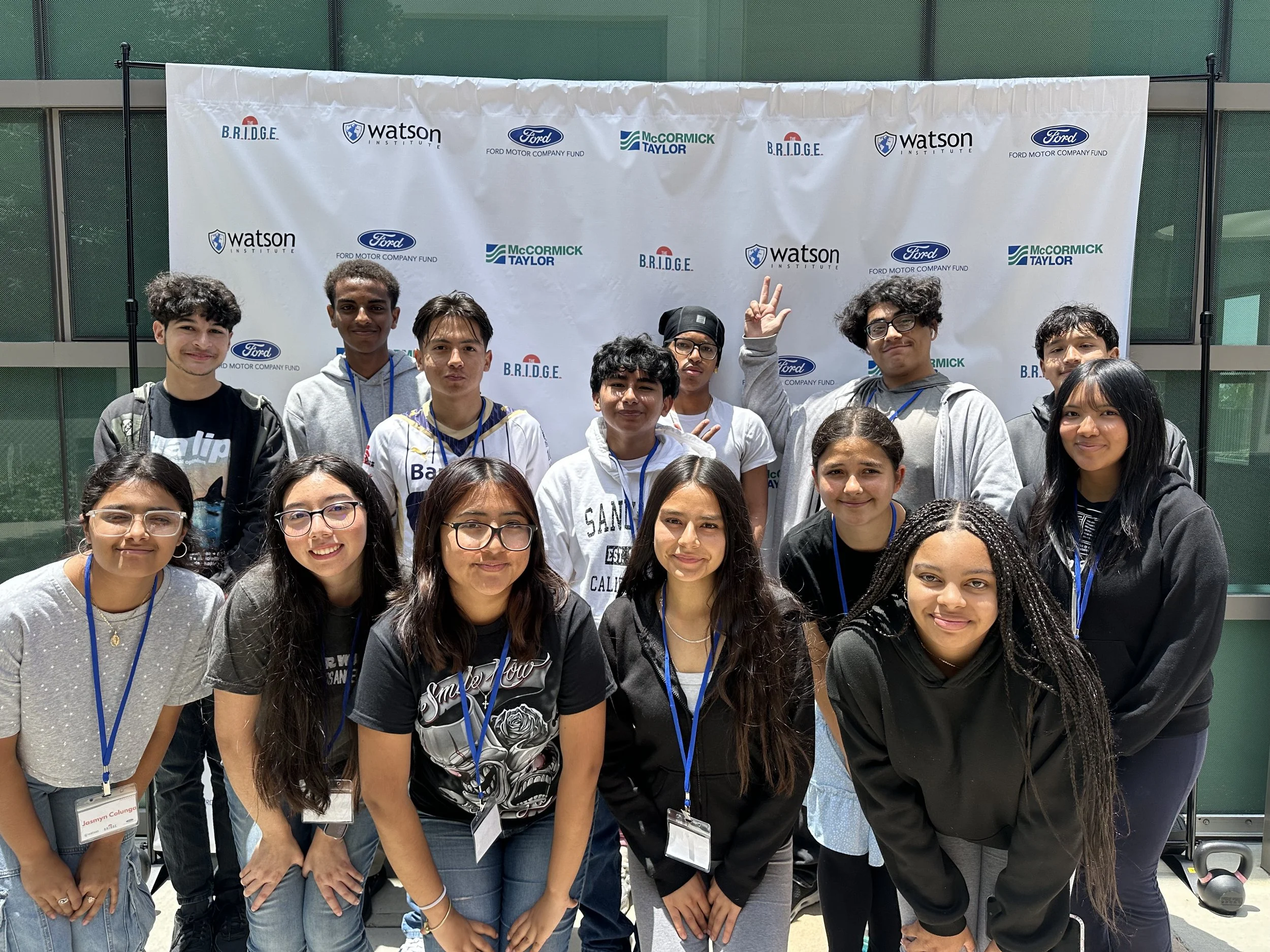 A diverse group of youth standing and smiling together in front of a BRIDGE and partner organization backdrop at a community event.
