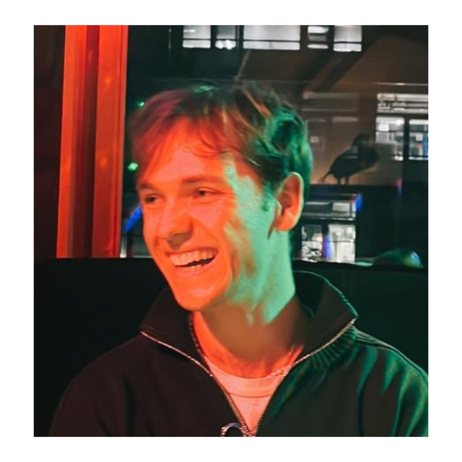 A young man with short brown hair, smiling in red lighting, looking to the side