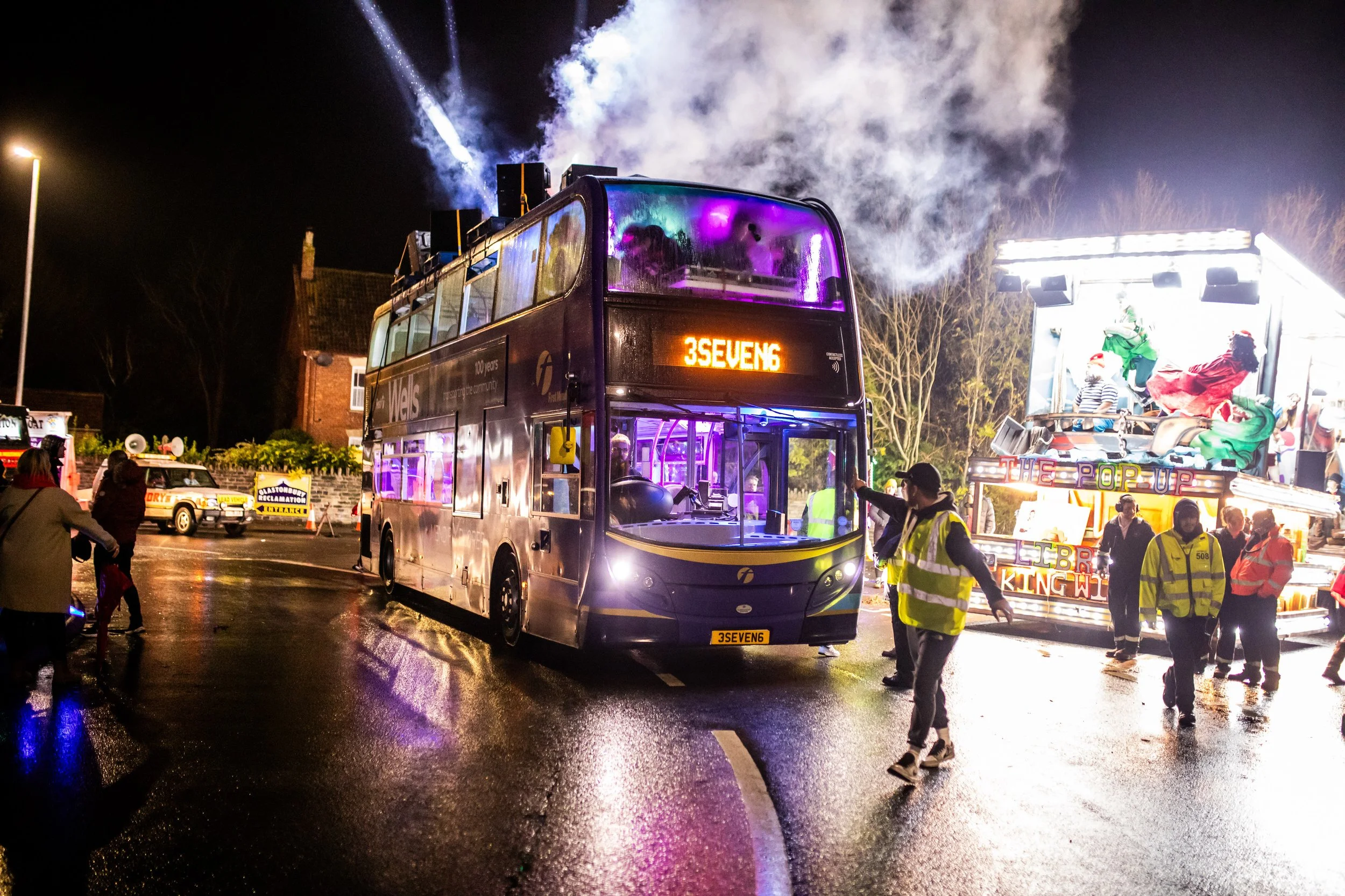 Nighttime scene of a crowded street with a double-decker bus, parade float, and people celebrating, some wearing Santa hats. The street is wet, reflecting colorful lights from the parade.