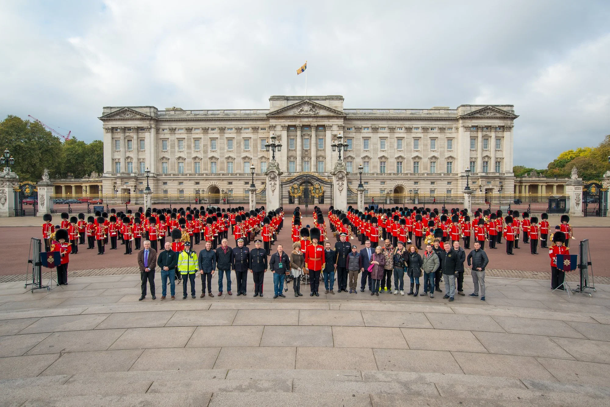 The exterior of Buckingham Palace with a large group of people, including guards in red uniforms and tall bearskin hats, standing in formation in front of the palace gates. The palace is a grand white building with columns and a flag flying on top, with a cloudy sky overhead.