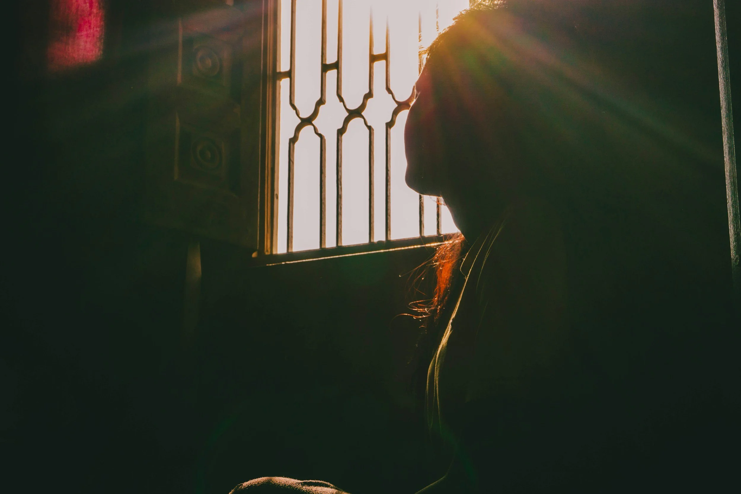 A person standing behind a window with bars during sunset, with sunlight creating a glare and lens flare in the image.