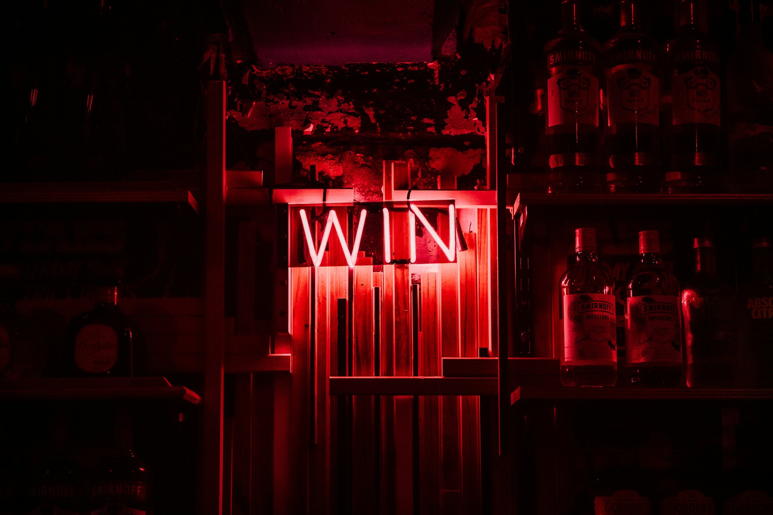 Red neon sign that says 'WIN' hanging on a wood wall inside a bar with shelves of liquor bottles around.