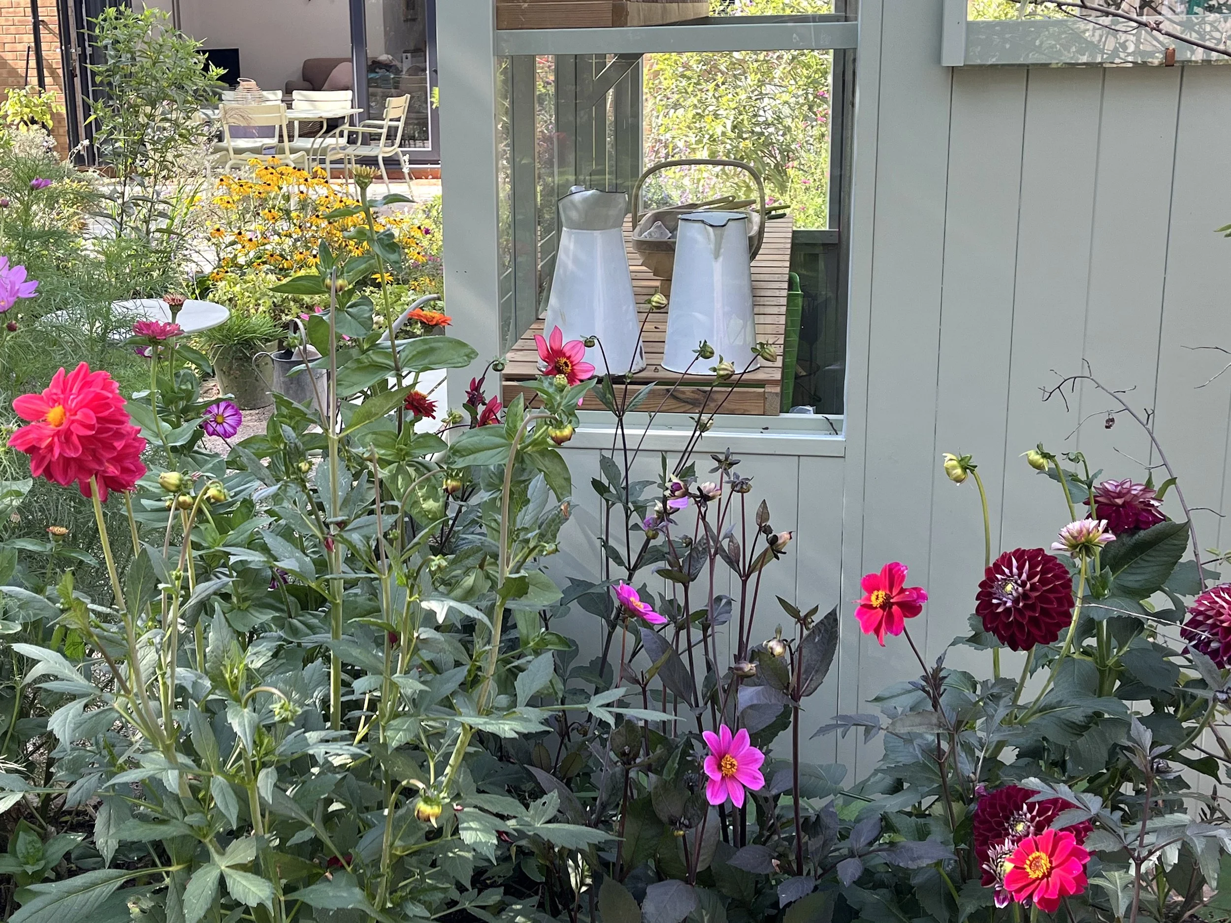 View of a garden with colorful flowers in front of a house with a window, showing a patio with chairs, a table, and two white watering cans on a wooden surface.