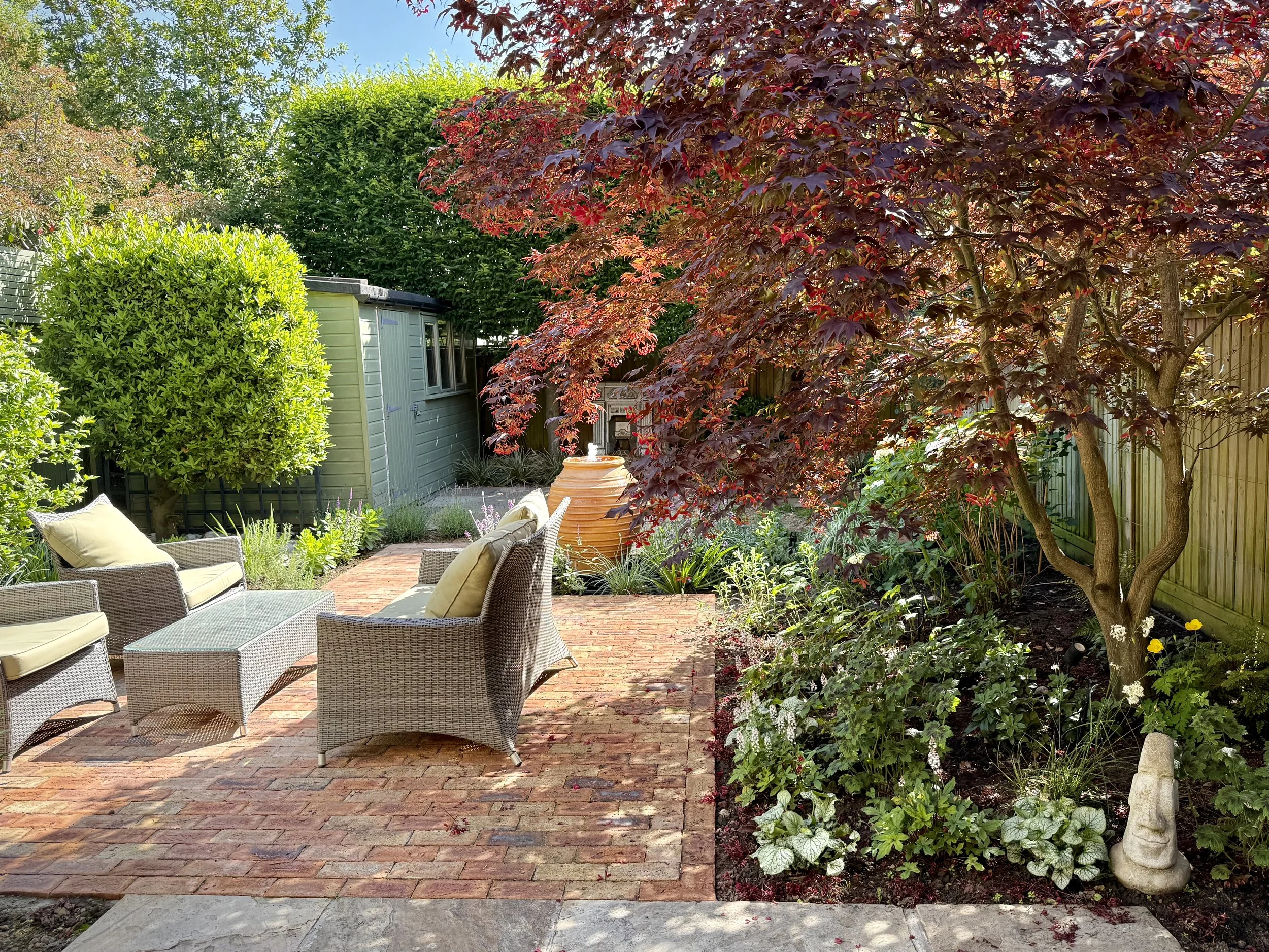 A garden terrace with brick flooring, beige outdoor chairs, surrounded by lush green and reddish trees, decorative plants, and garden ornaments.