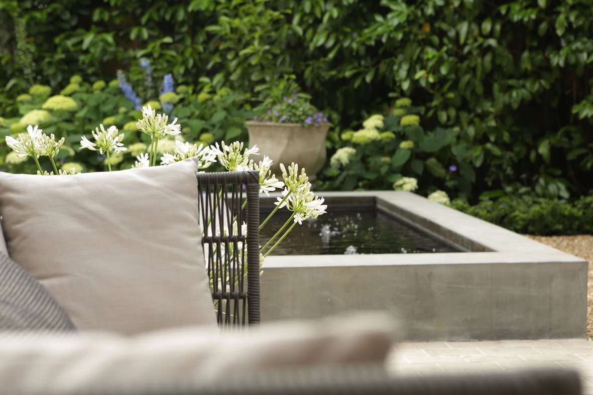 Outdoor garden with a modern rectangular water feature, surrounded by green foliage and blooming white flowers, with a seating area including pillows.