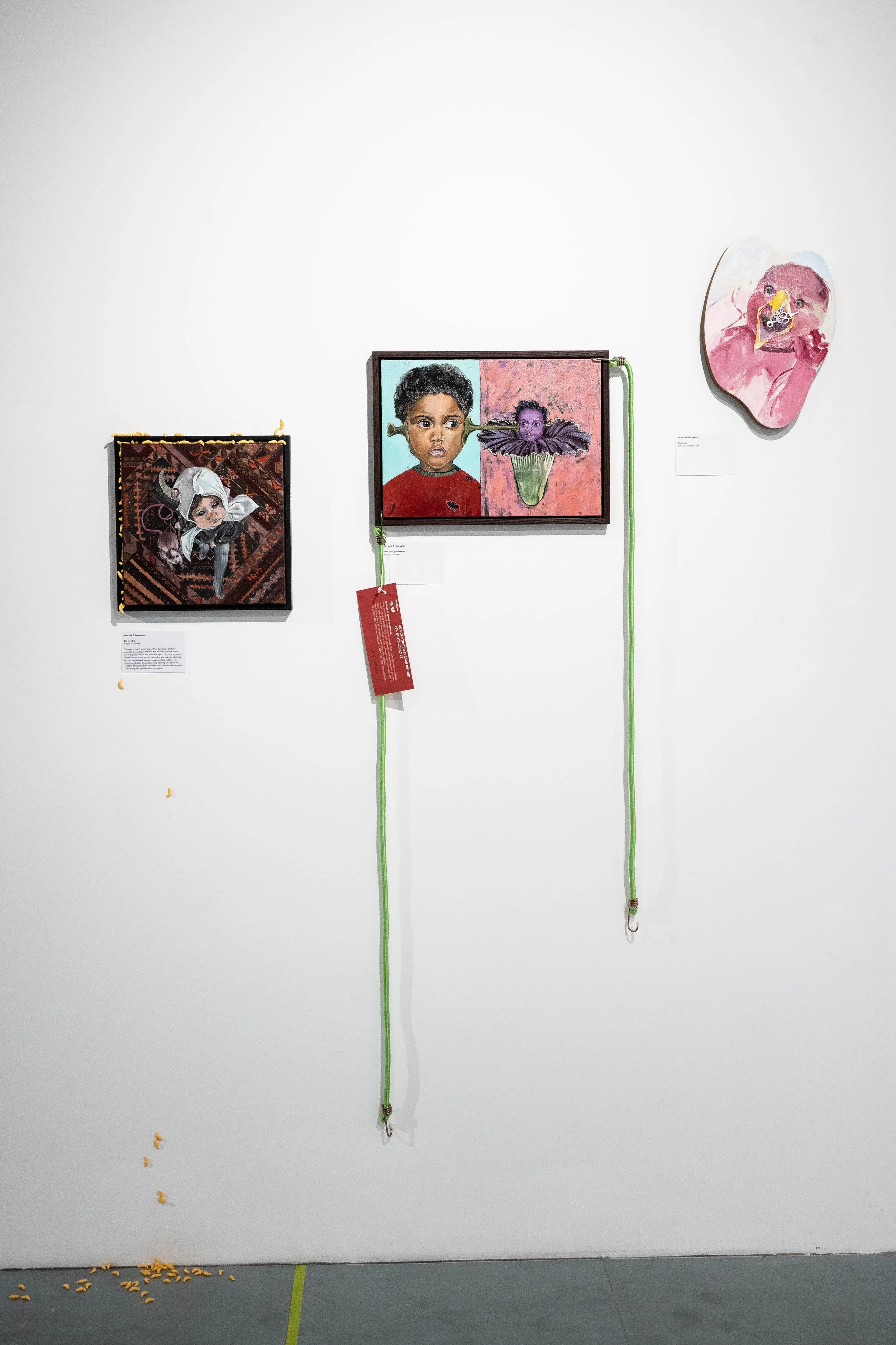 An art exhibit featuring three mixed media portrait artworks on a white gallery wall. Two green cords hang from the wall, attached to the second artwork. The left artwork depicts a young girl with cat ears and a bow. The middle artwork shows a boy wi