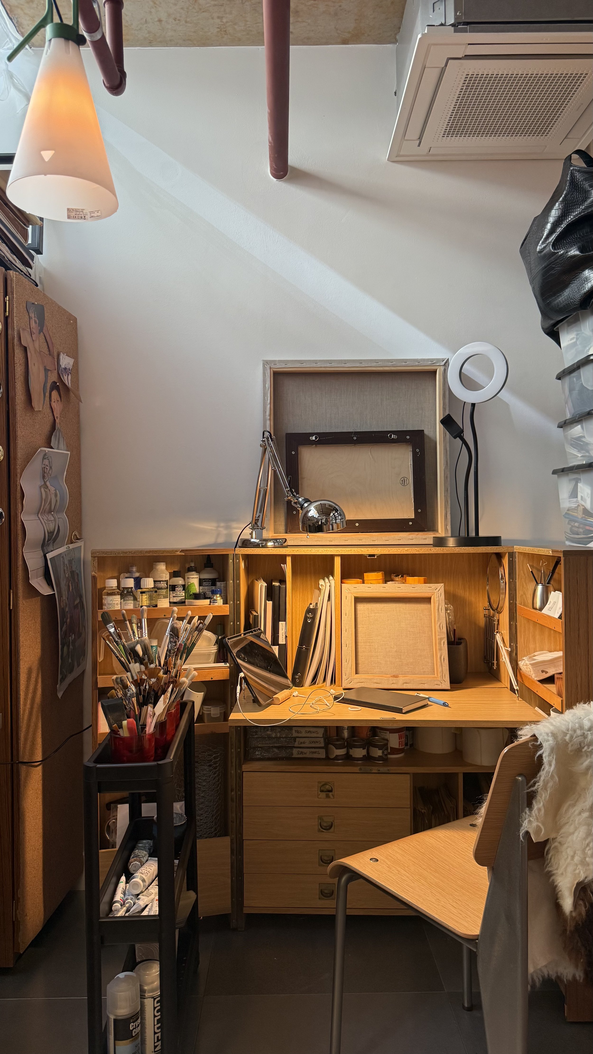A small workspace with a wooden desk, shelves filled with art supplies, a desk lamp, and a chair with a sheepskin throw. The wall has photos and art pinned up, and there is a rolling cart with markers and other art tools. A black bag hangs on the right side, and a ceiling vent and pipes are visible above.