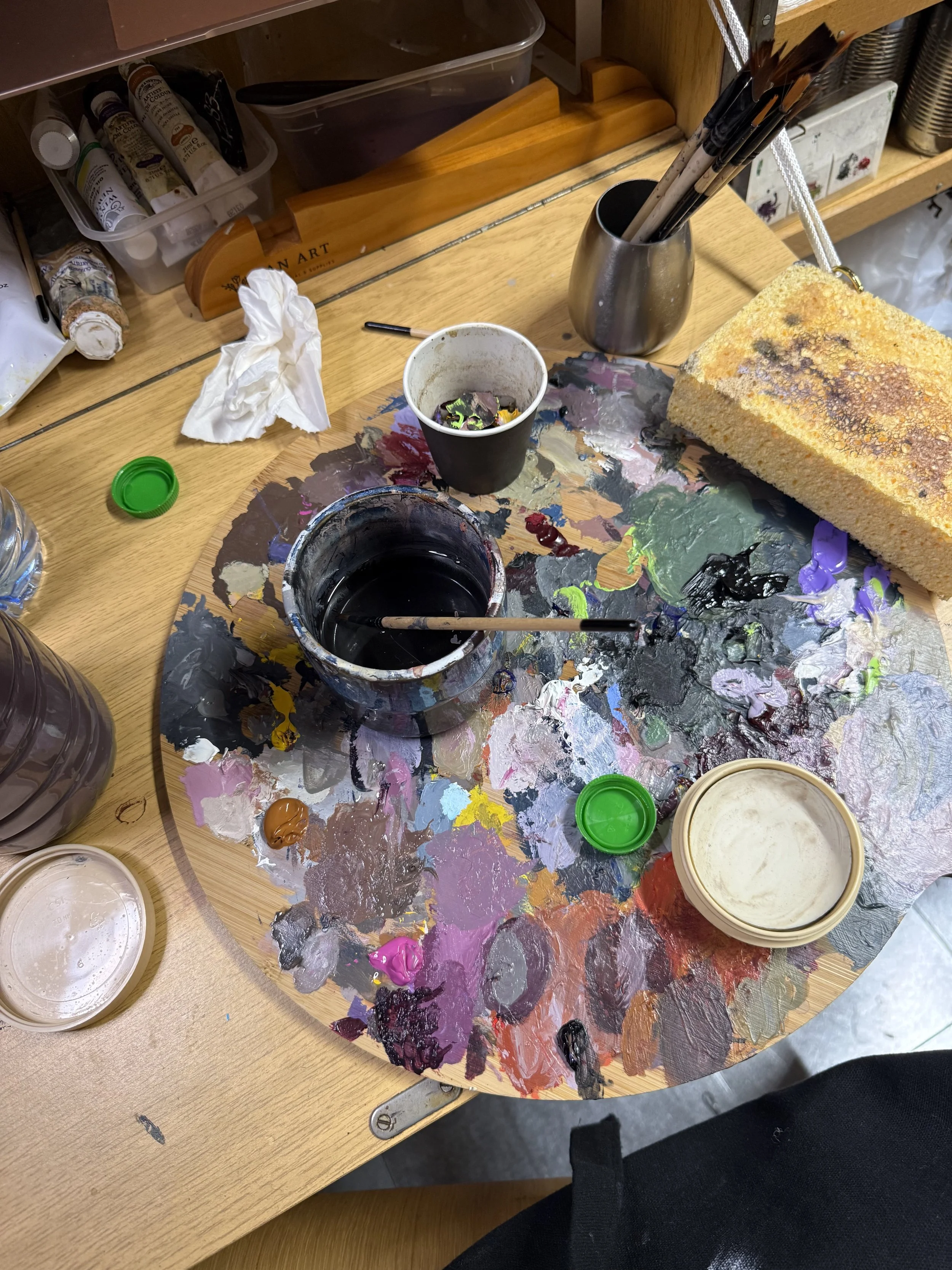 An artist's workspace with a wooden palette covered with a mixture of vibrant paint colors, an open container of black paint, various paint tubes, a crumpled tissue, and a sponge with yellow-orange paint. There are also brushes in a metallic holder, a glass of water, and open containers of other paint colors on a wooden table.