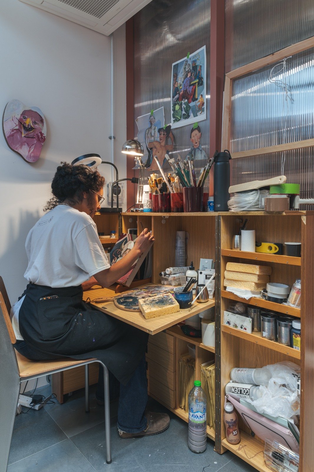 An artist working on a painting in a cozy art studio. The workspace is filled with art supplies, paintings on the wall, and shelves with supplies and tools.