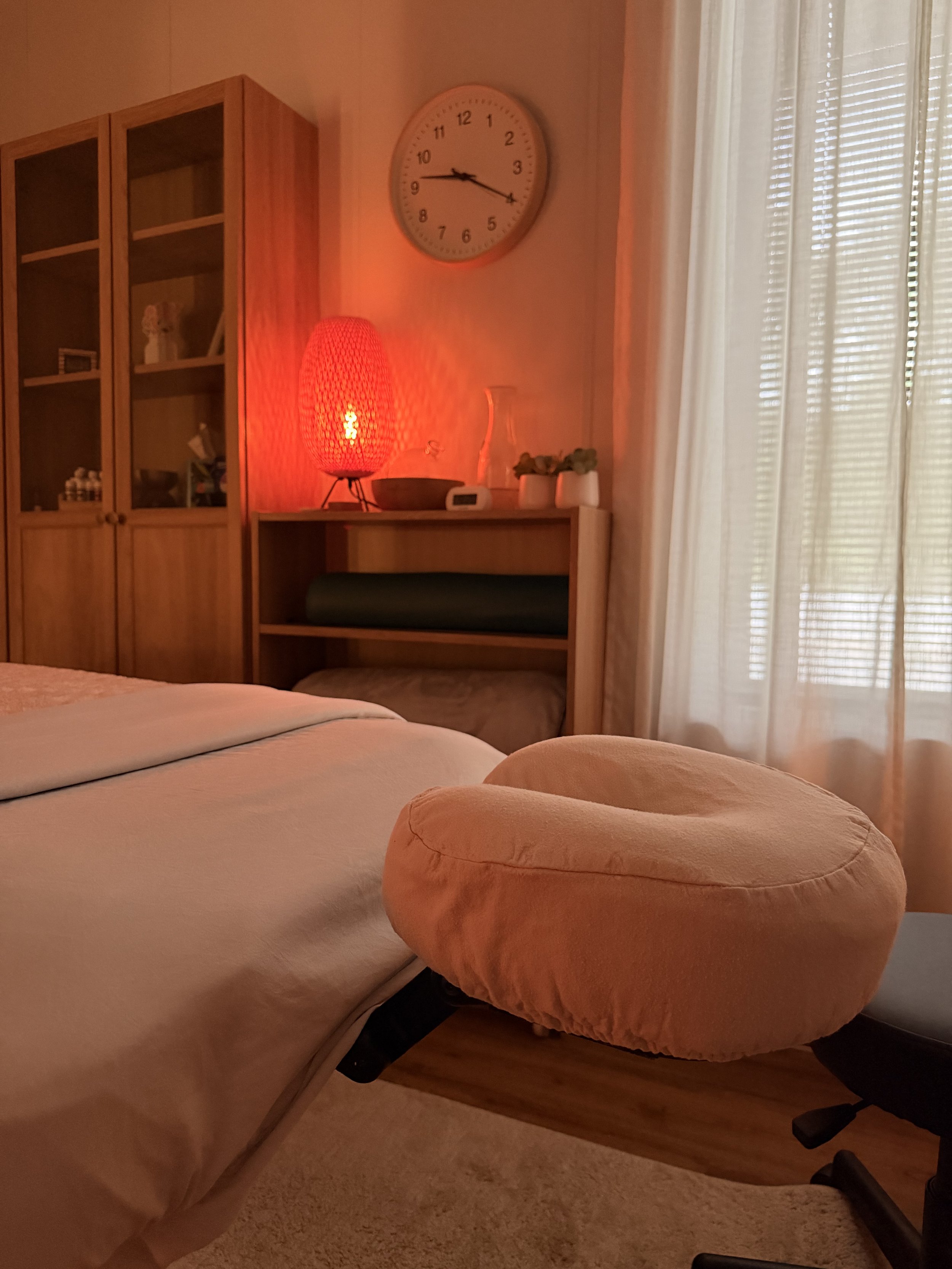 A cozy bedroom corner with a bed, a soft pink round pillow on a beige carpet, a wooden bookcase with glass doors, a red bedside lamp emitting a warm glow, a white ceramic vase, and two small potted plants. A wall clock shows the time as 9:43, and curtains allow natural light into the room.