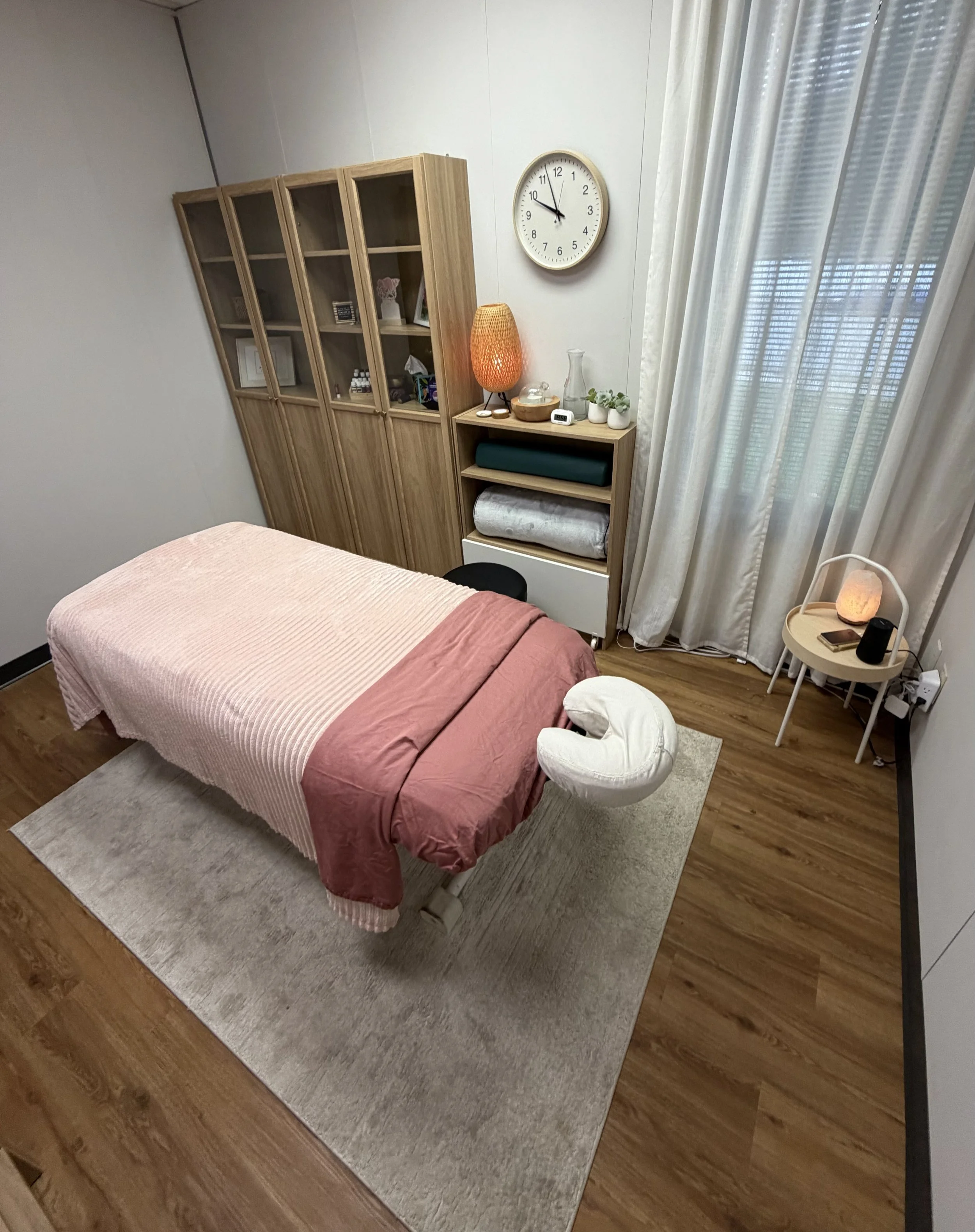 A massage treatment room with a massage table covered in a pink bedspread, a wooden bookshelf, a small table with a lamp, a clock on the wall, and a window with curtains.