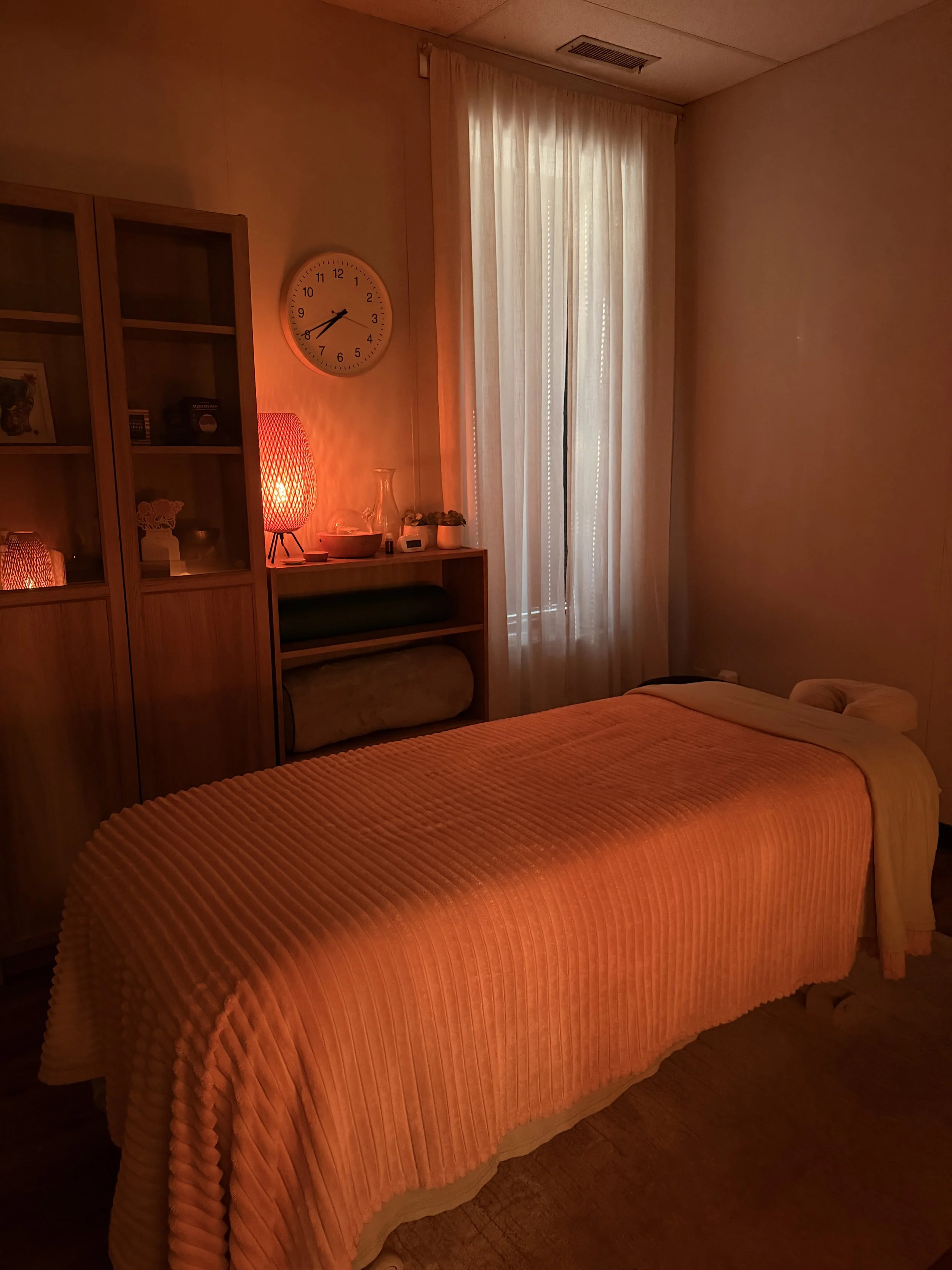 Be Well Massage Studio Greensboro NC relaxing massage therapy room with warm amber lighting, massage table with orange blanket, sheer curtains, and spa decor