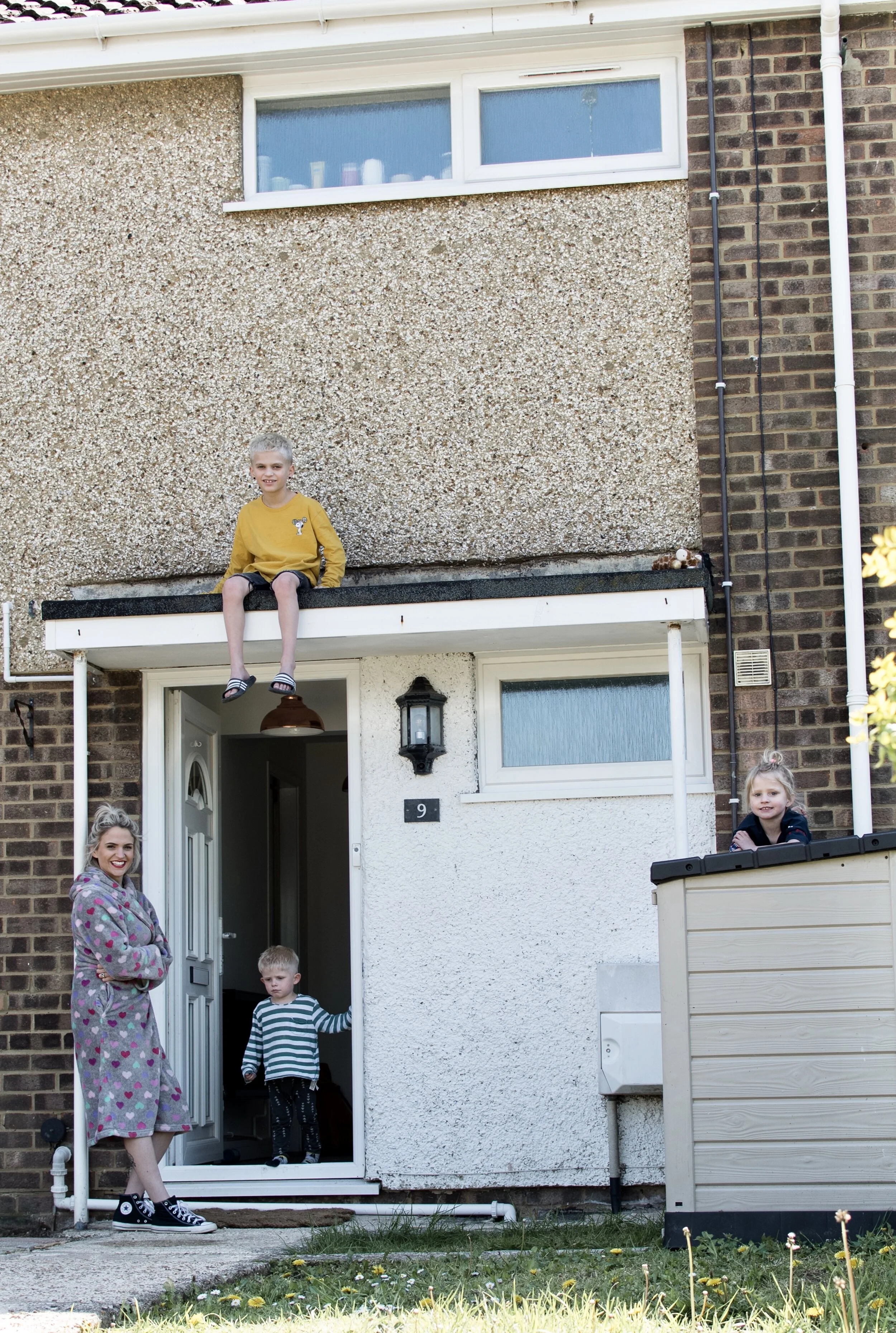 Family in front of brick house, child sits on porch roof, door open, other children nearby, person in robe standing.