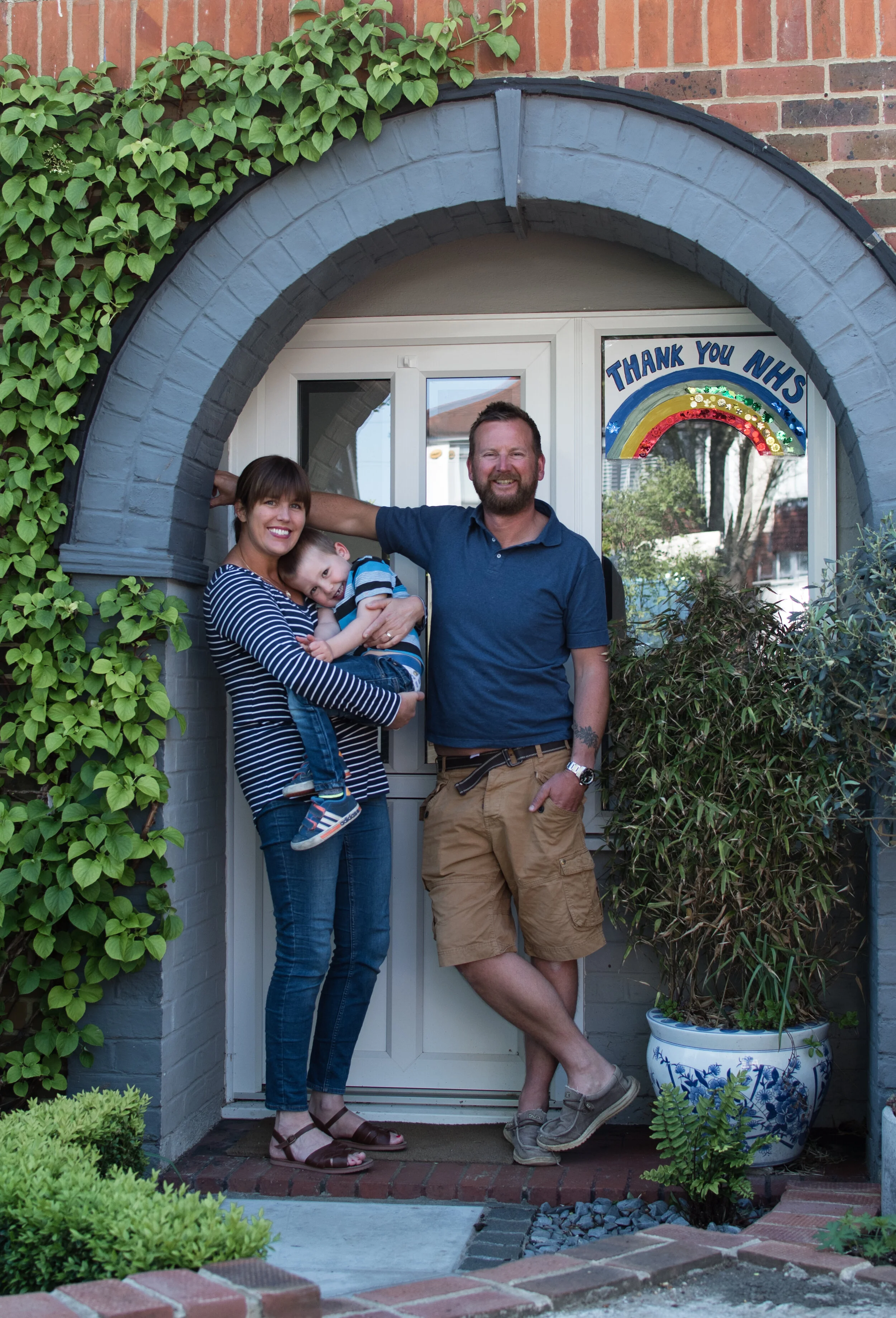 A family of three stands in front of a doorway. The mother holds a young child, and the father leans against the door frame. A "Thank you NHS" sign with a rainbow is displayed on the door. There are plants nearby, including ivy and a potted plant.