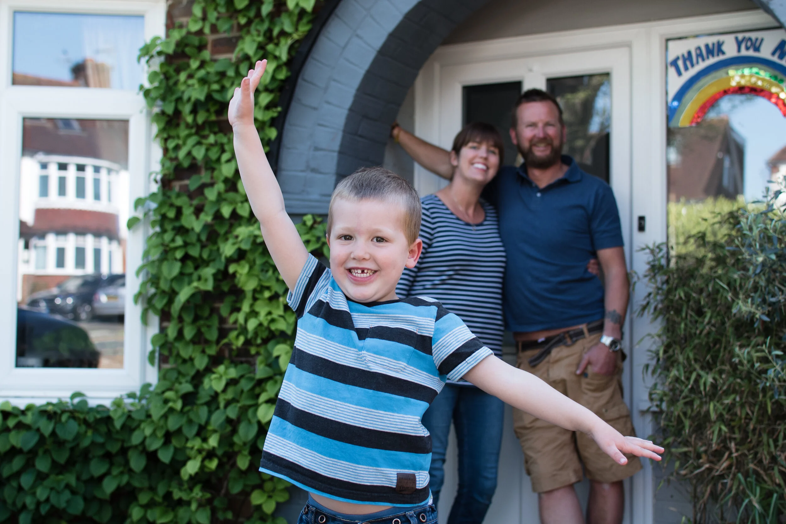 A cheerful boy with short hair in a blue and black striped shirt raises his arms happily in front of a smiling couple by their doorway. The house has green plants climbing the wall and a sign with a children's drawing of a rainbow and a message of th