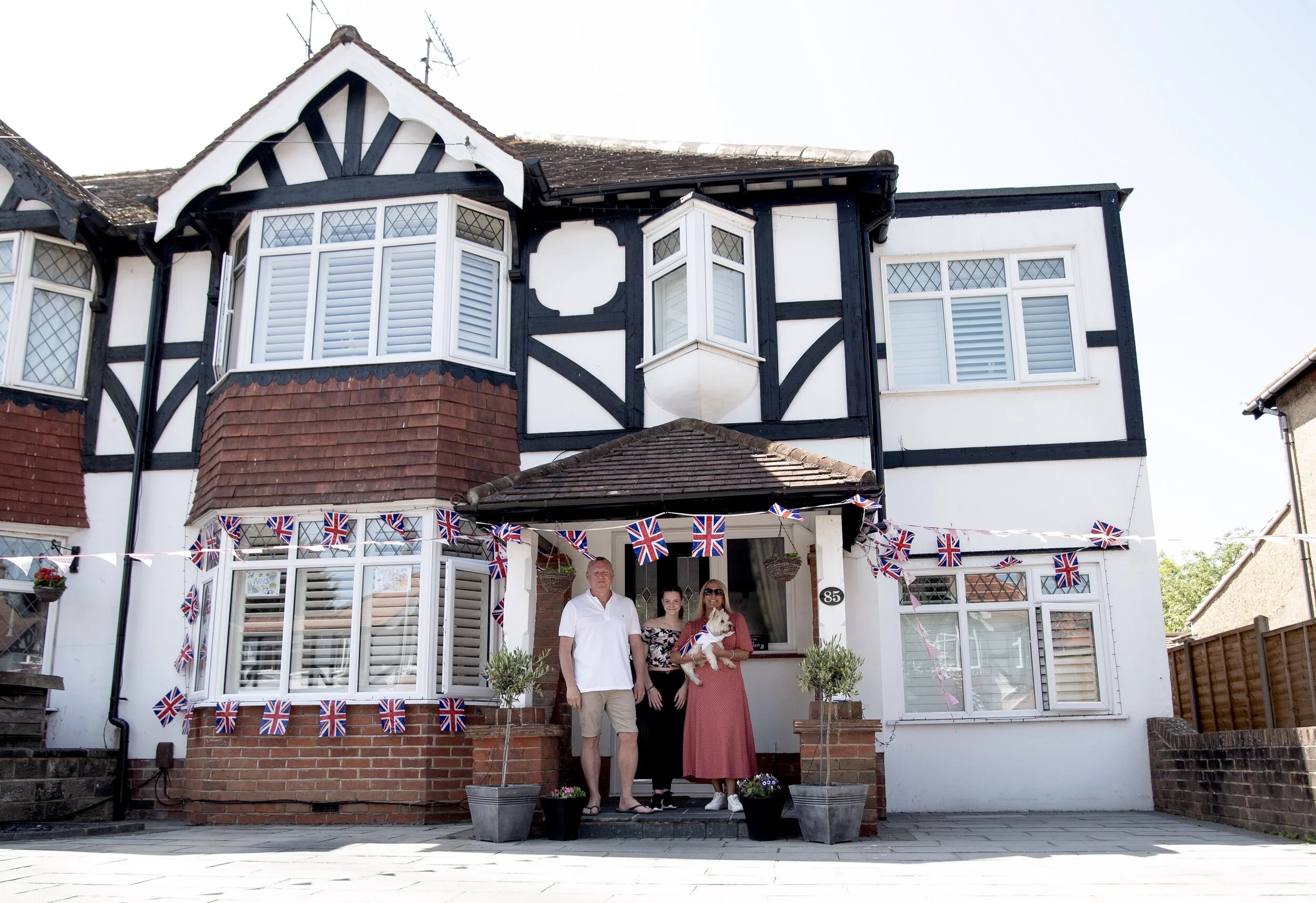 House decorated with Union Jack flags, three people and a dog in front, green potted plants by the entrance.