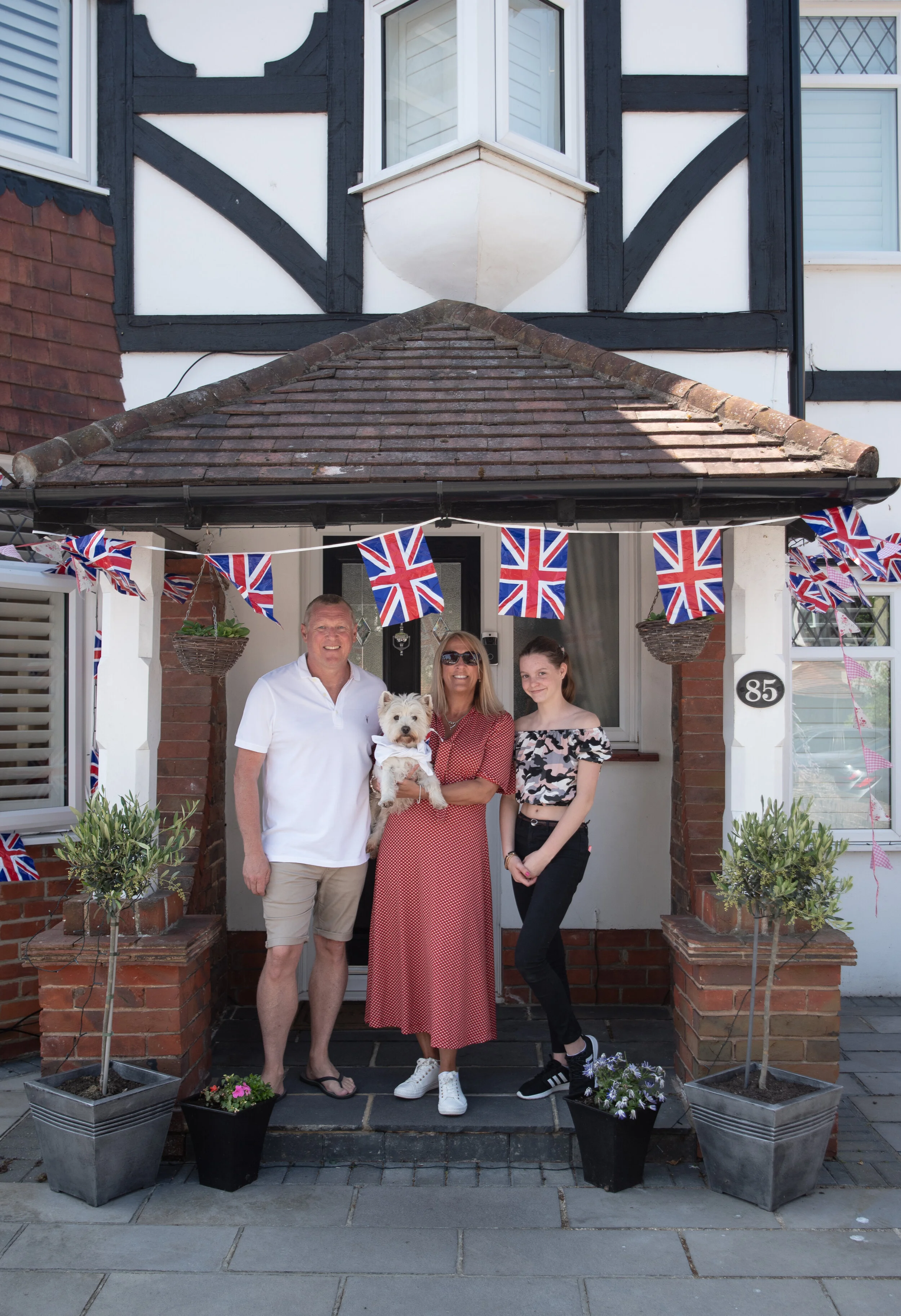 Family standing in front of house decorated with British flags, holding a small white dog.