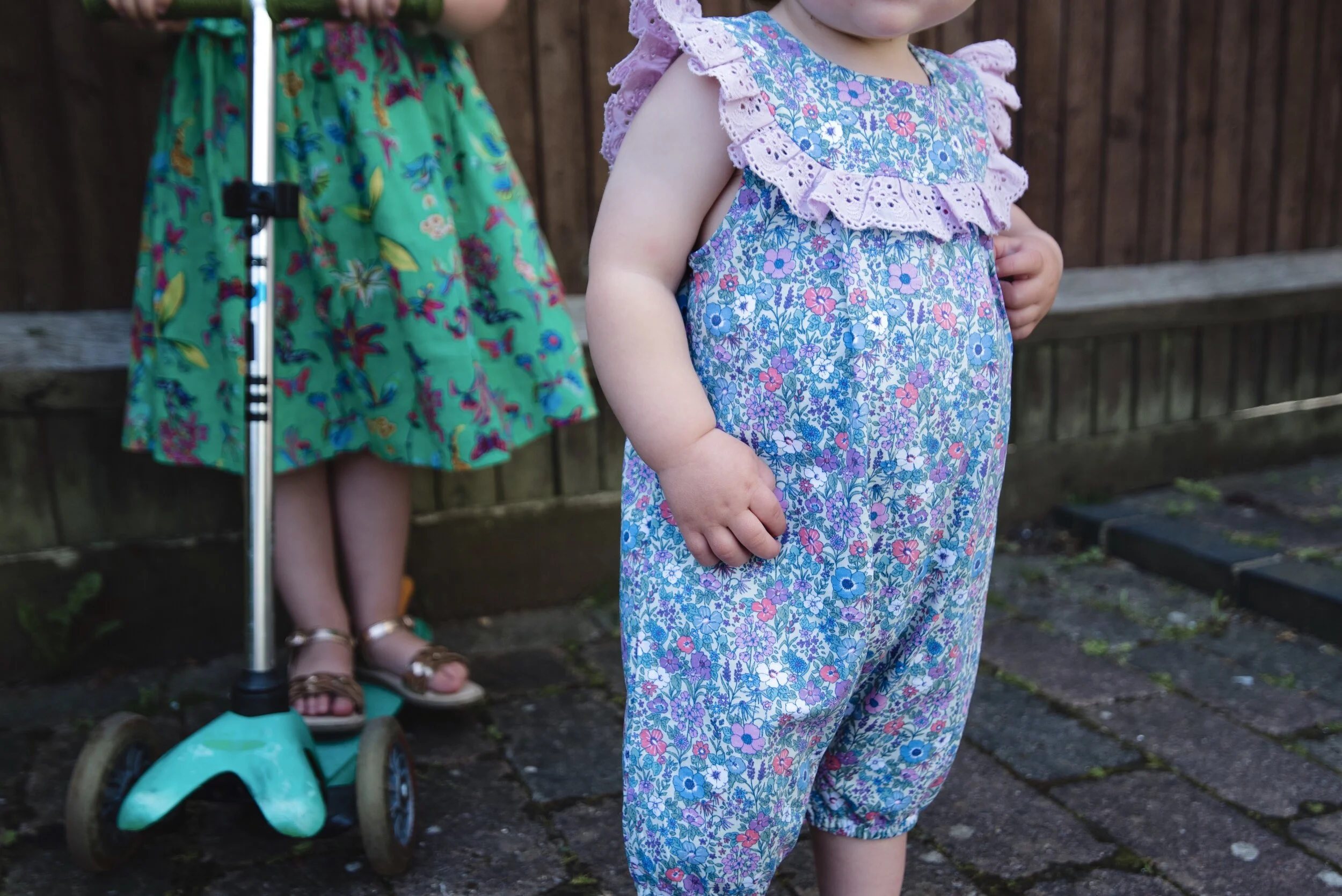 Two children in floral outfits; one in a green dress with a scooter, and another in a blue floral romper.