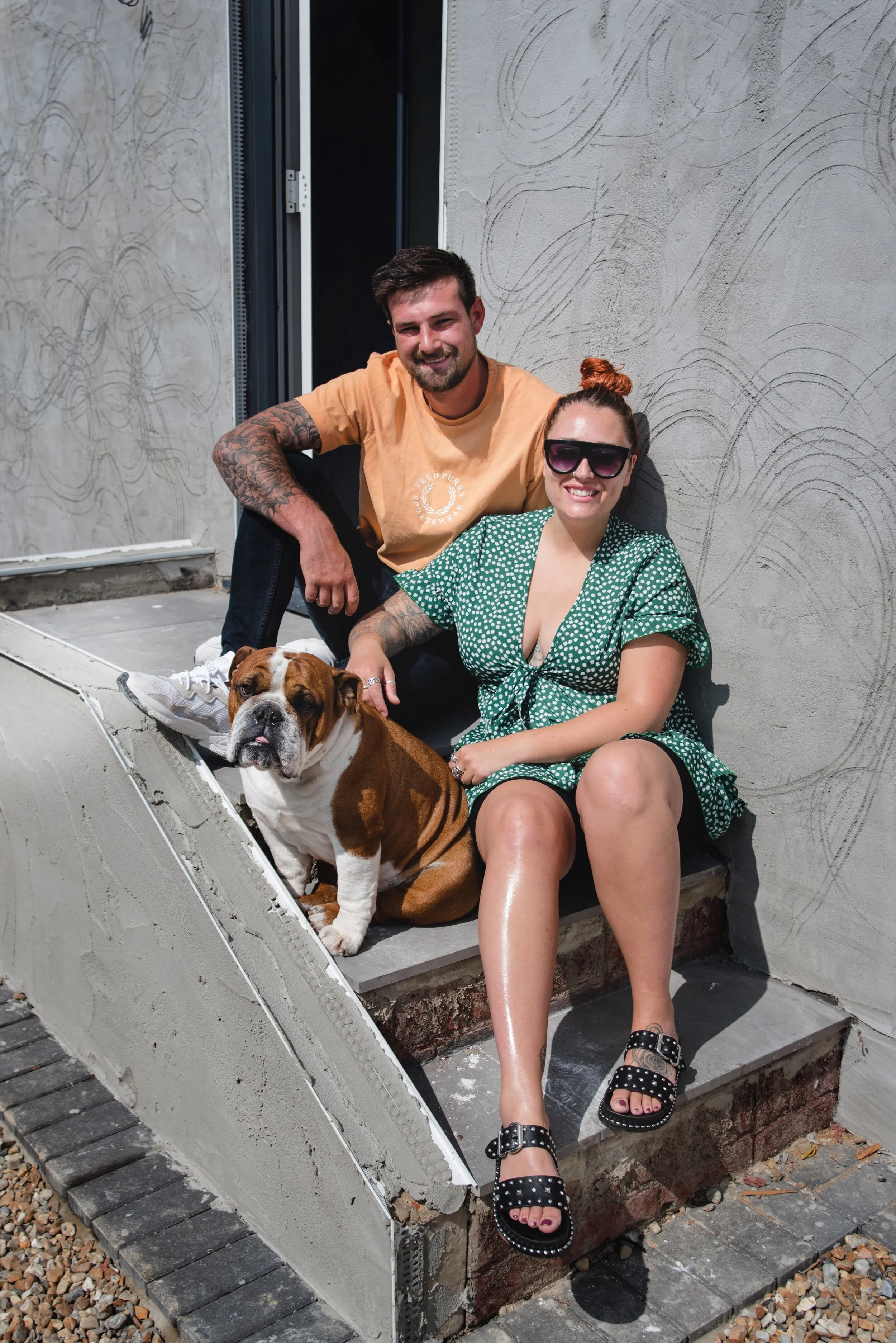 A man and a woman sitting on concrete steps with an English bulldog. The man is wearing a yellow T-shirt and has tattoos on his arms. The woman is wearing sunglasses and a green polka dot dress. The bulldog is sitting between them.