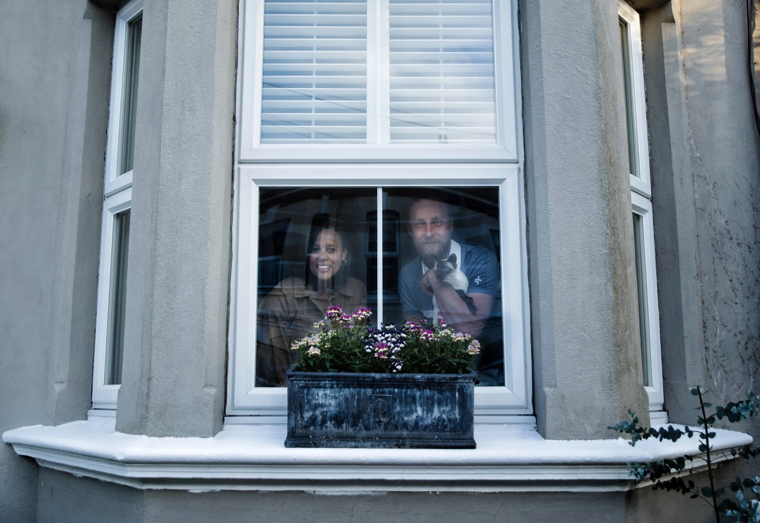 Two people and a cat looking out of a window with potted flowers on the sill.