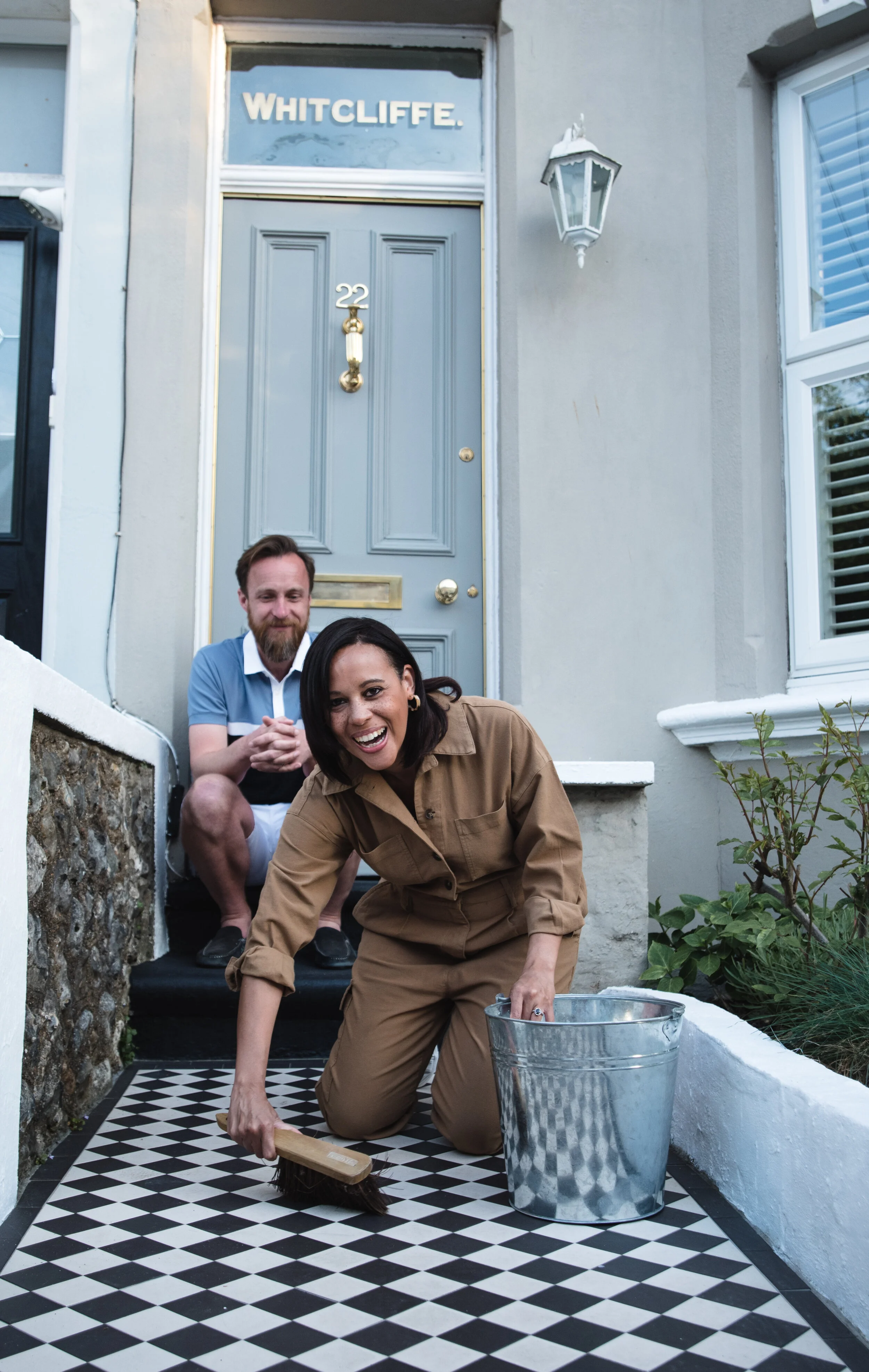 Two people on a porch; a woman kneeling and cleaning the checkered floor with a brush, bucket nearby, a man sitting behind her."}