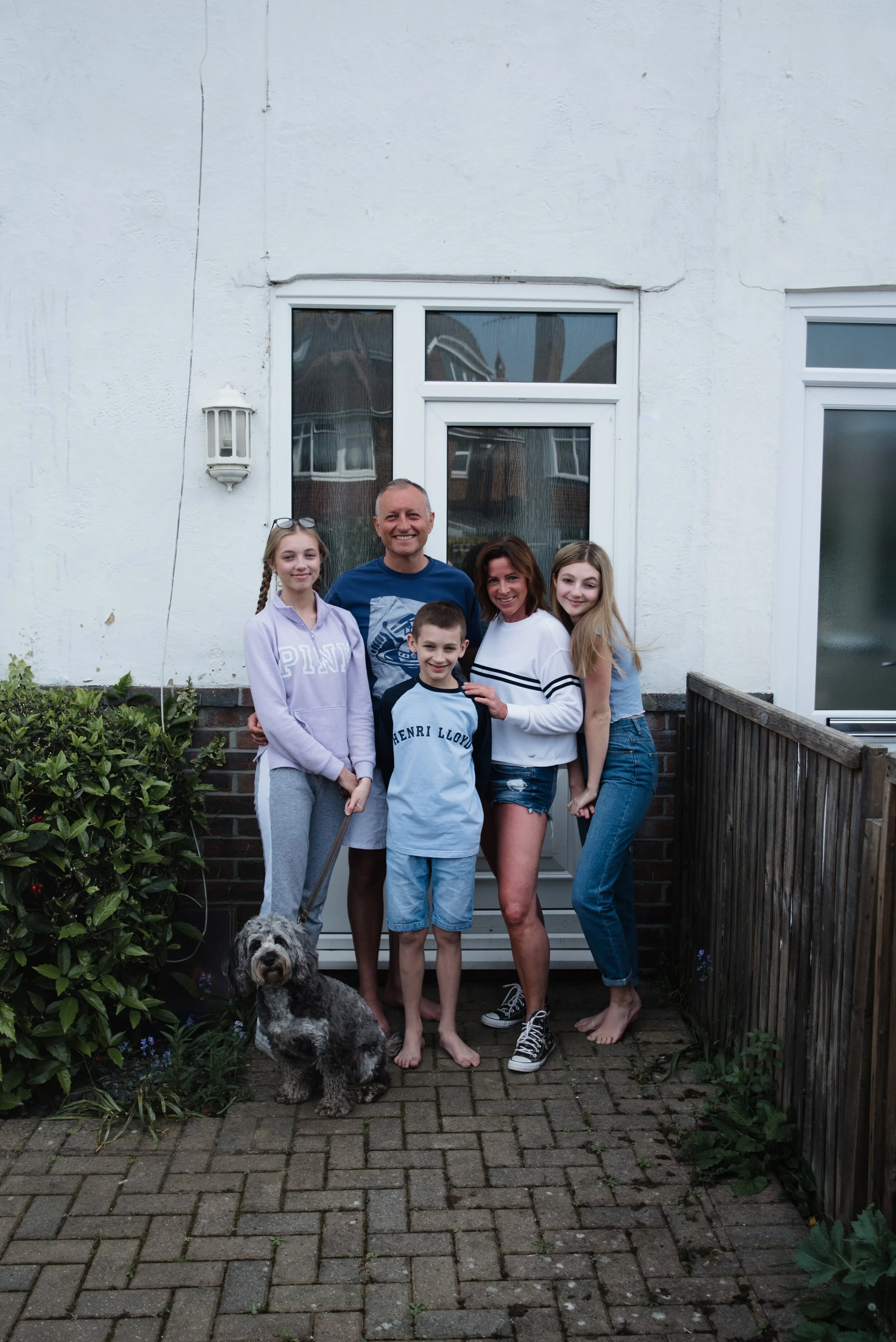 Family of five with a dog posing outside a house with white walls, including two men, two women, one girl, and a dog on a leash.