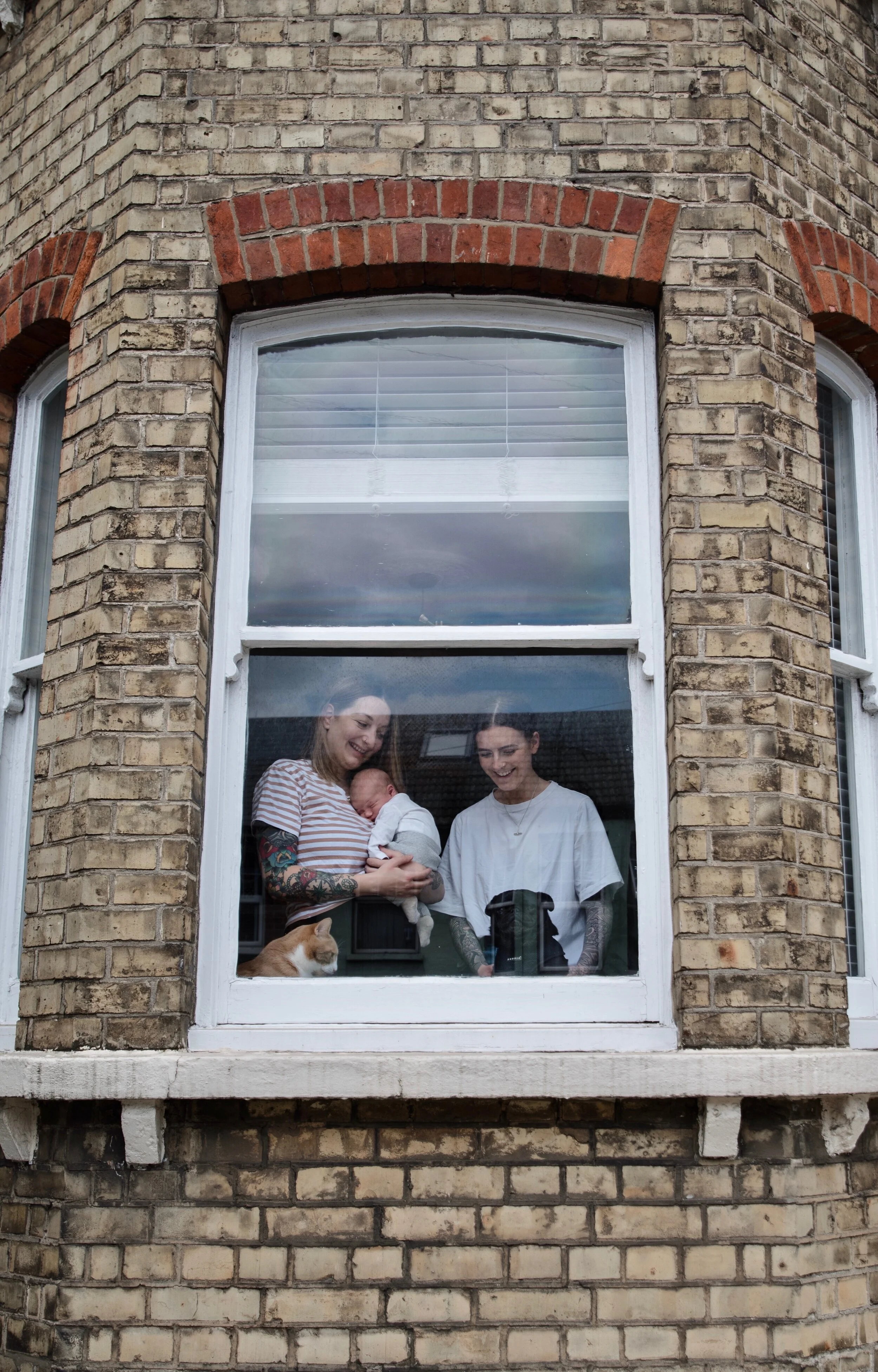 Two people inside a brick building looking out a window, one holding a baby. A cat sits on the windowsill.