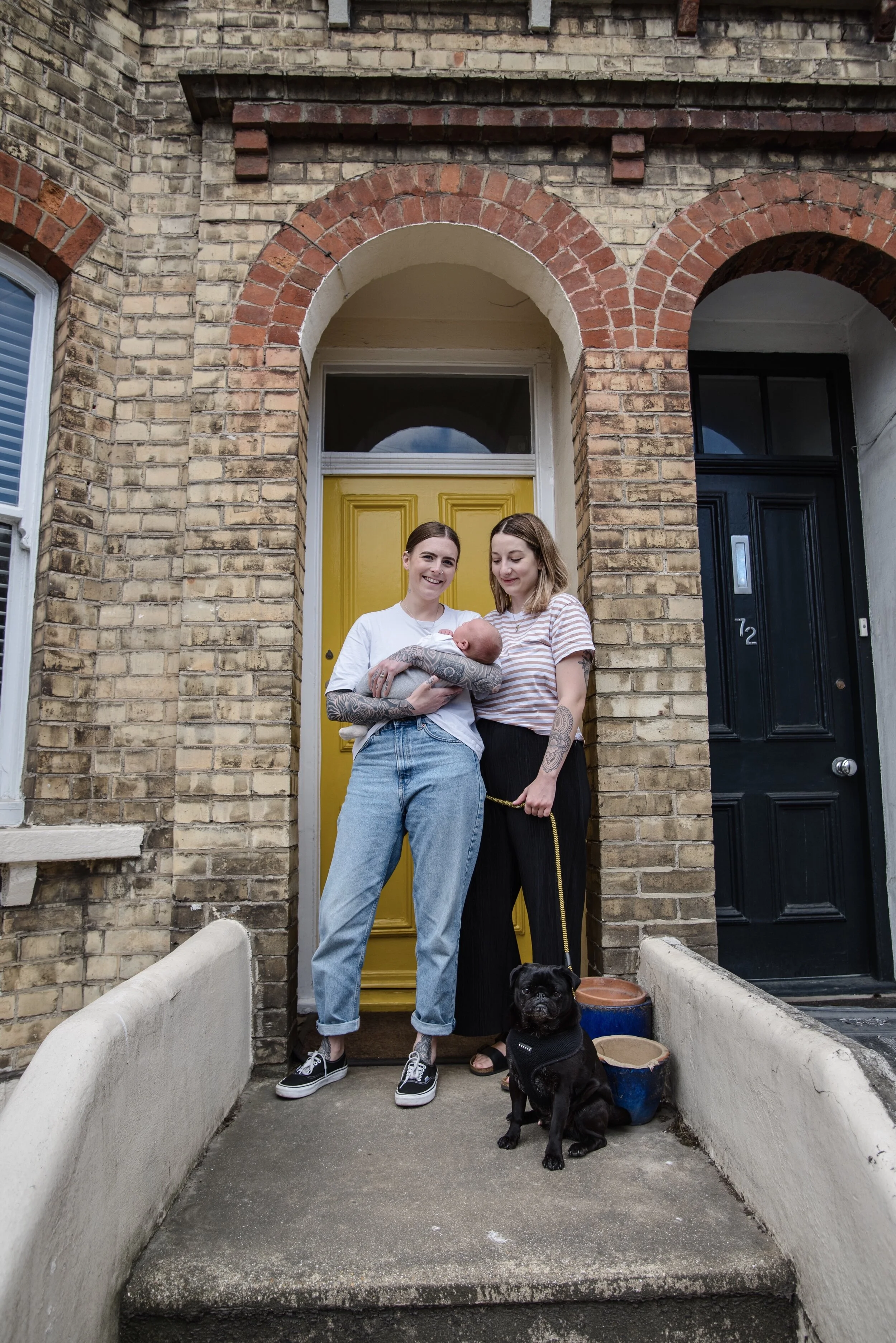 Two people standing on the steps of a brick house with a yellow door, one holding a baby. A black dog sits nearby. There are potted plants on the right.