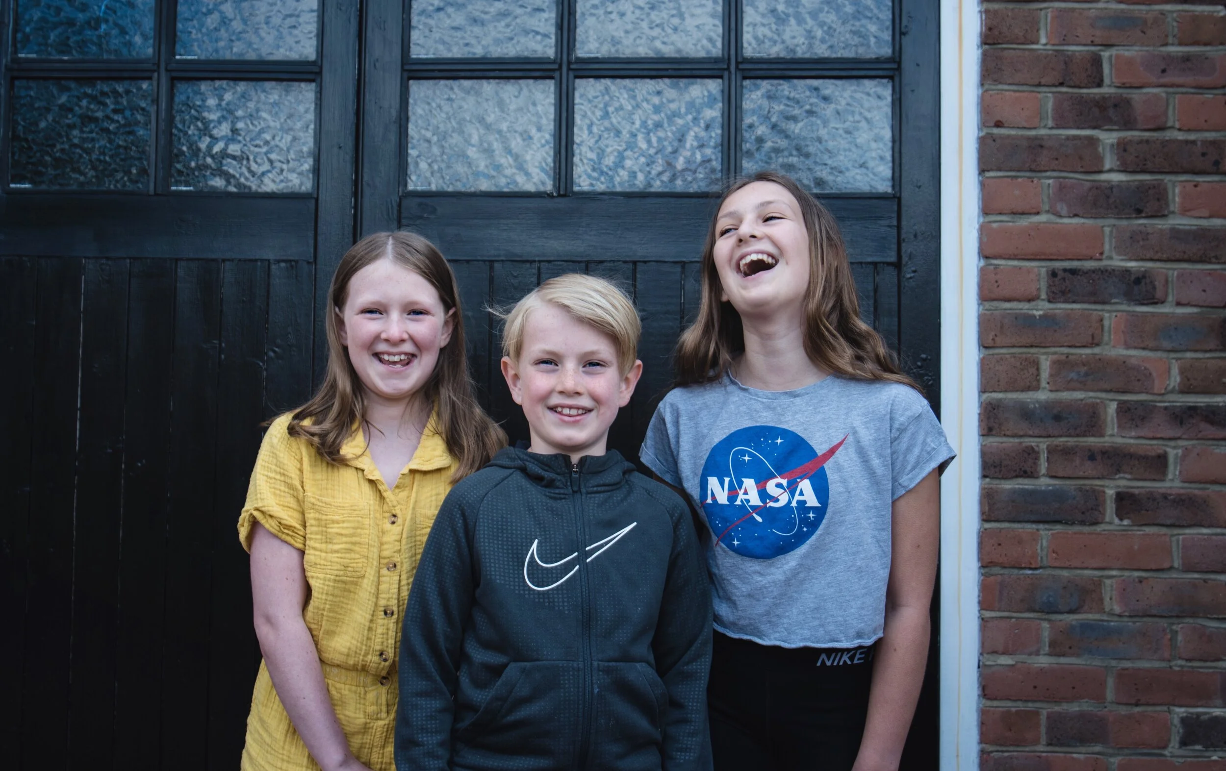 Three children smiling in front of a black door with glass panels. One wears a yellow shirt, another a gray Nike jacket, and the third a gray NASA T-shirt.