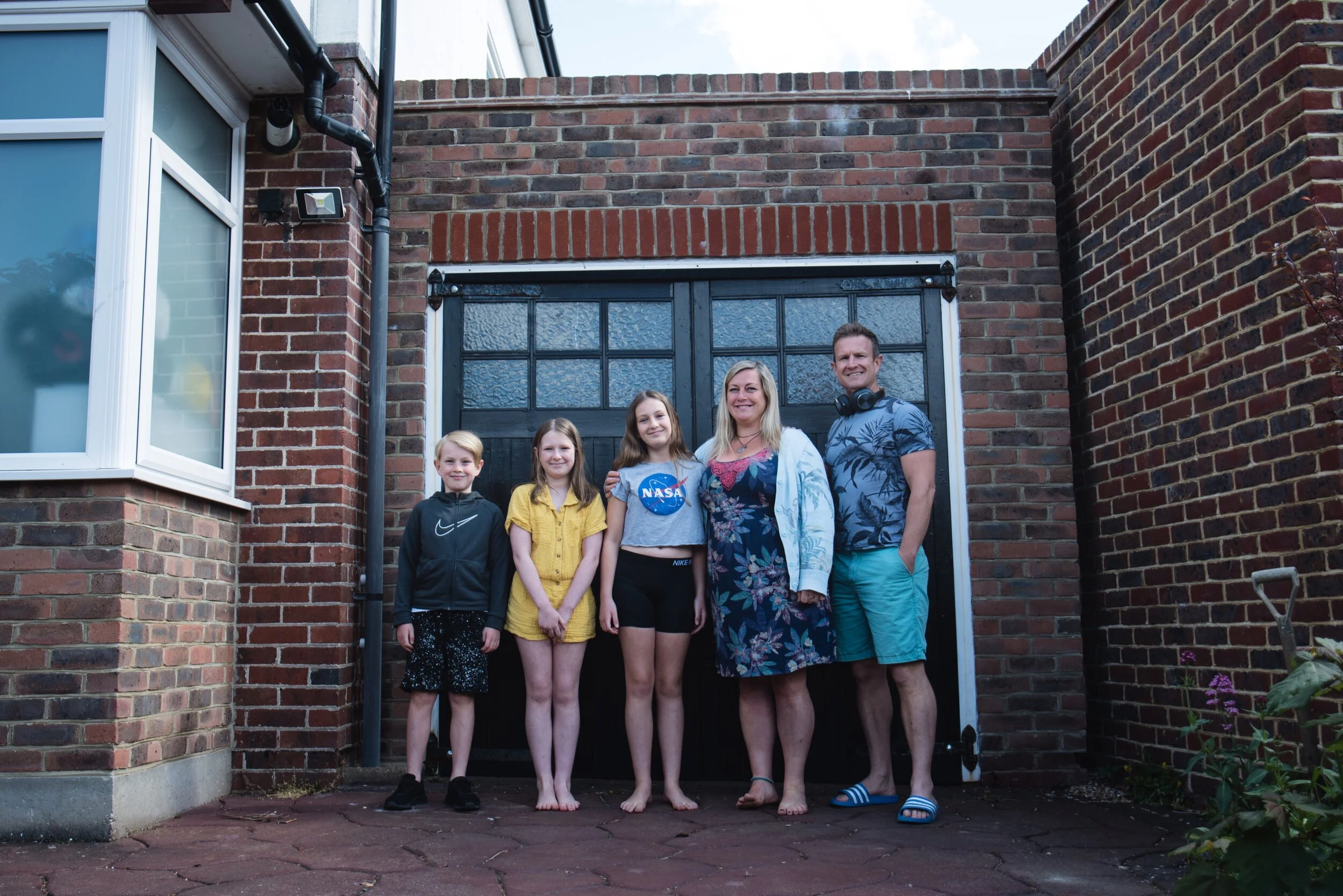 A family of five standing in front of a brick building. The group includes two adults and three children. The adults are on the right, and the children are on the left. They are standing on a paved area next to a large set of black doors with windows
