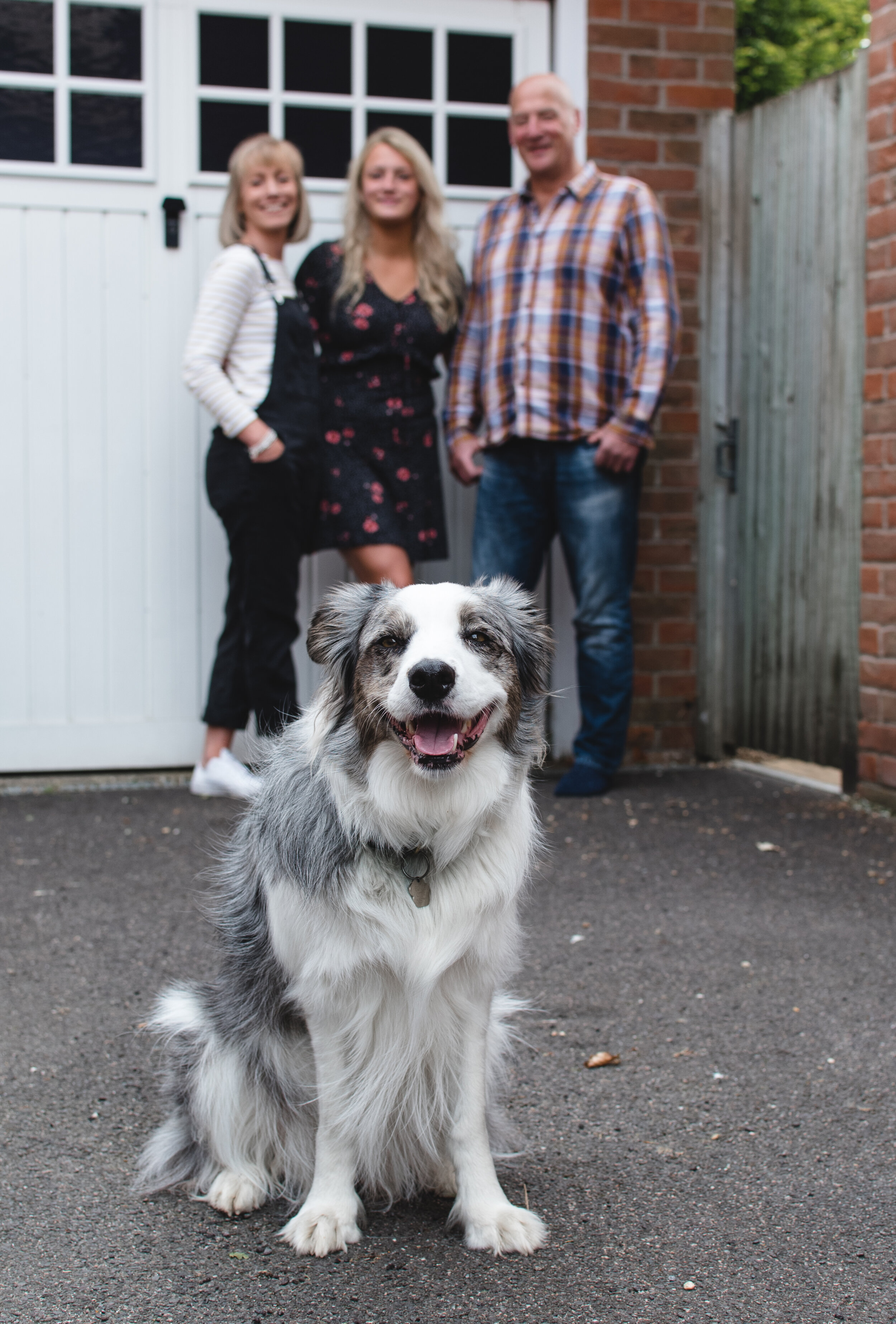 Smiling border collie with three people standing in the background outside a garage.