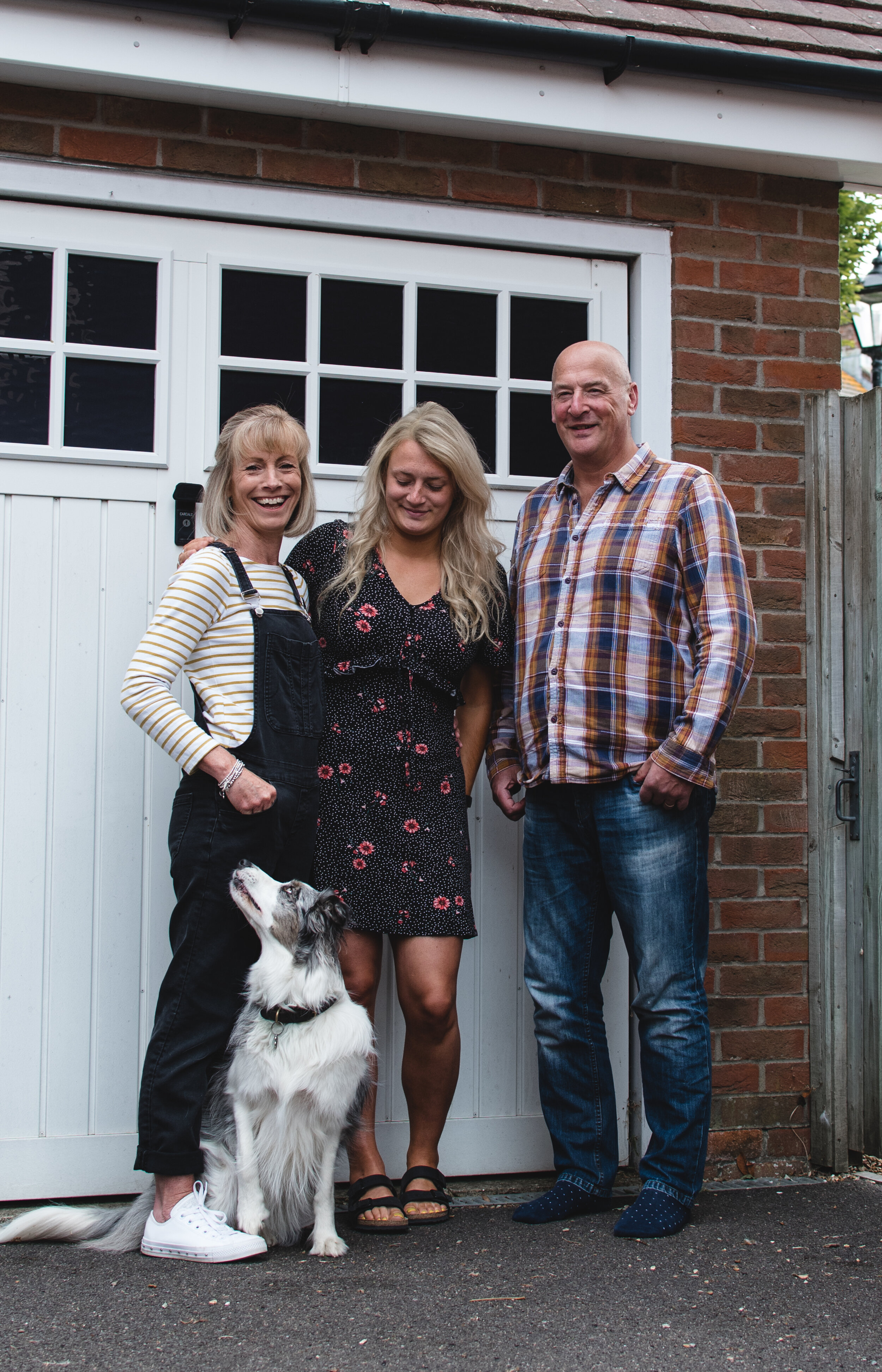 Three adults standing in front of a brick house, with white garage doors in the background. A dog is sitting at their feet, looking up at them.
