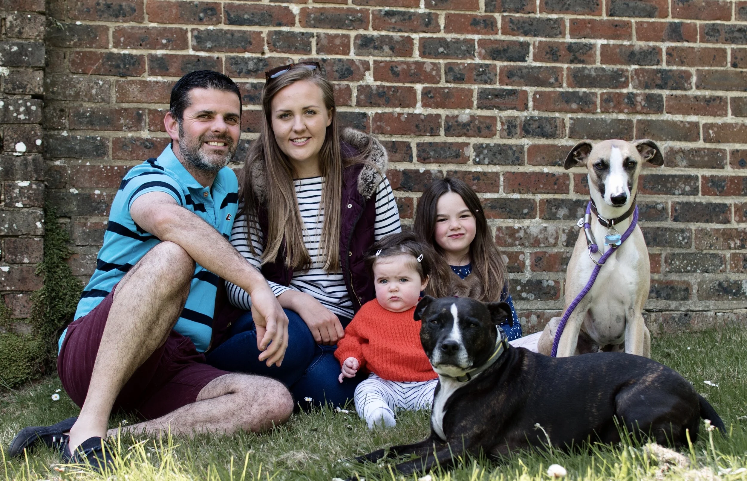 Family with two dogs sitting on grass in front of brick wall.