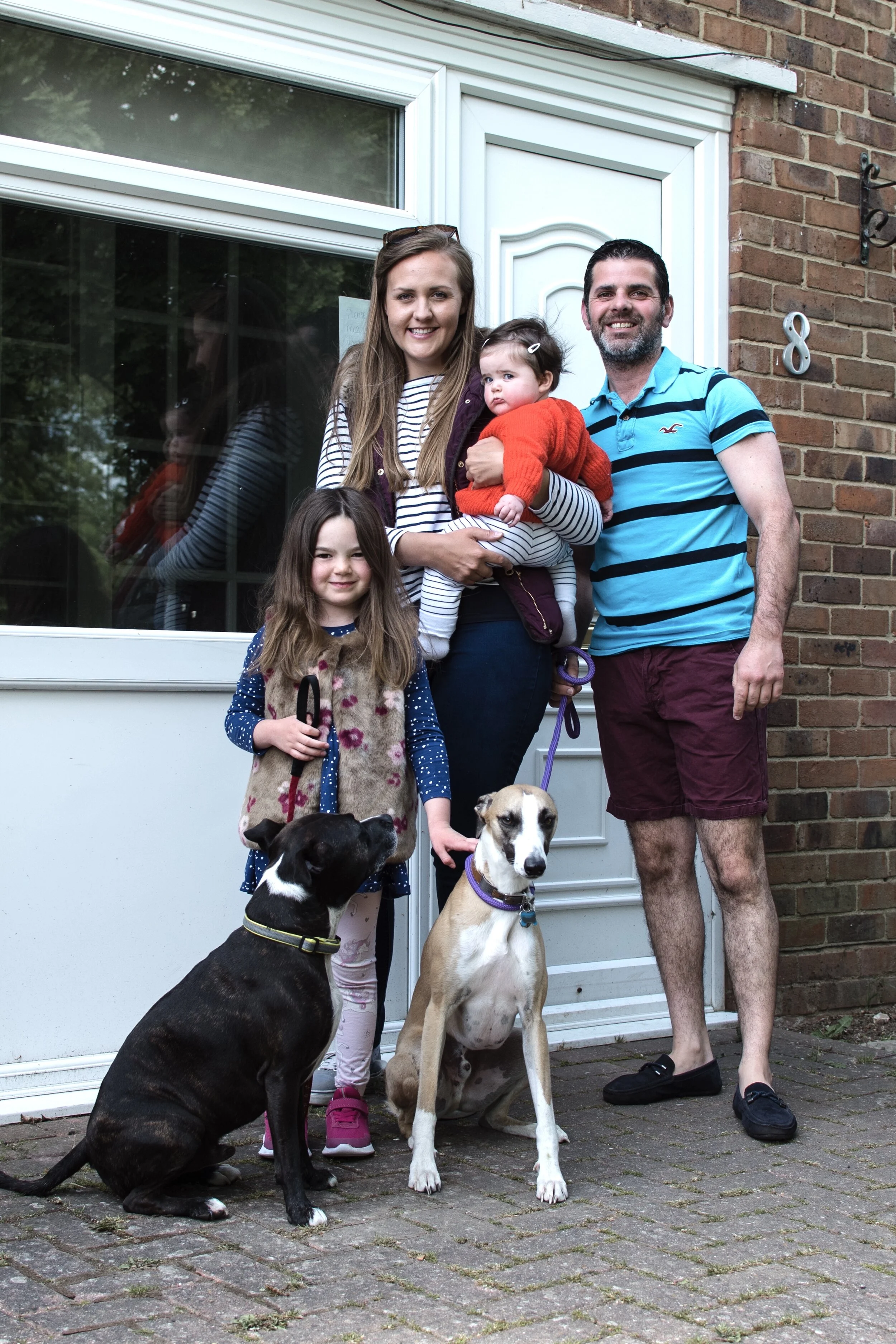 Family with two children and two dogs standing in front of a house with a white door and brick wall.