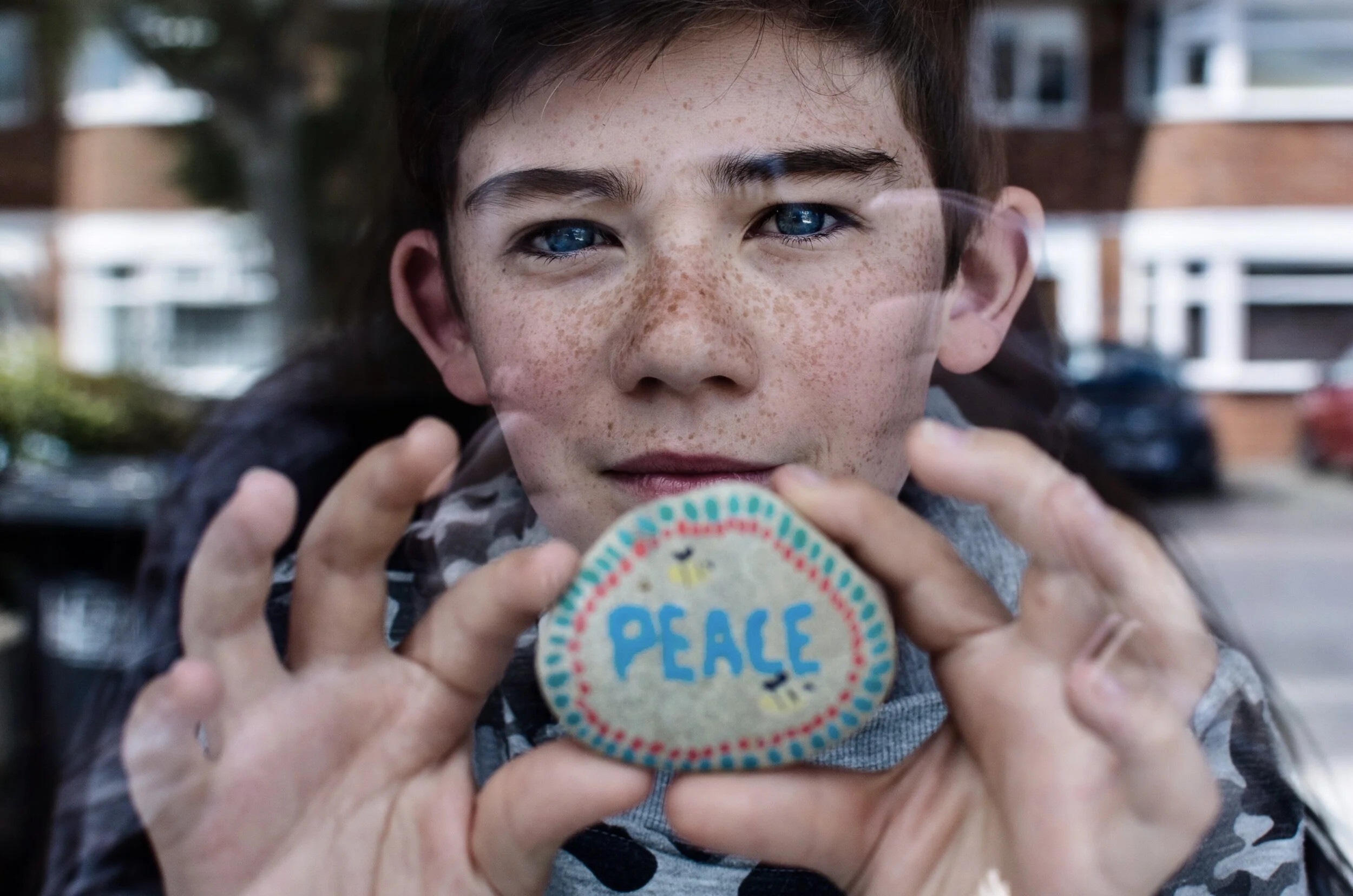 Young boy holding a painted rock with the word "Peace" written on it, visible through a glass window.