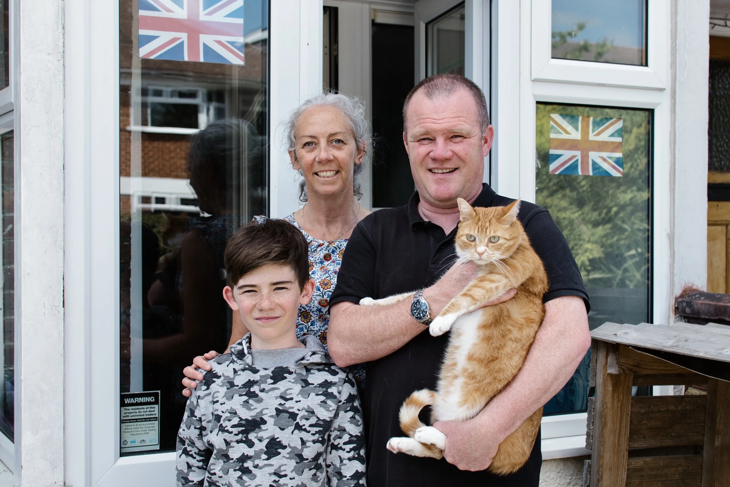 Family of three with a cat in front of a house with British flags in the windows.