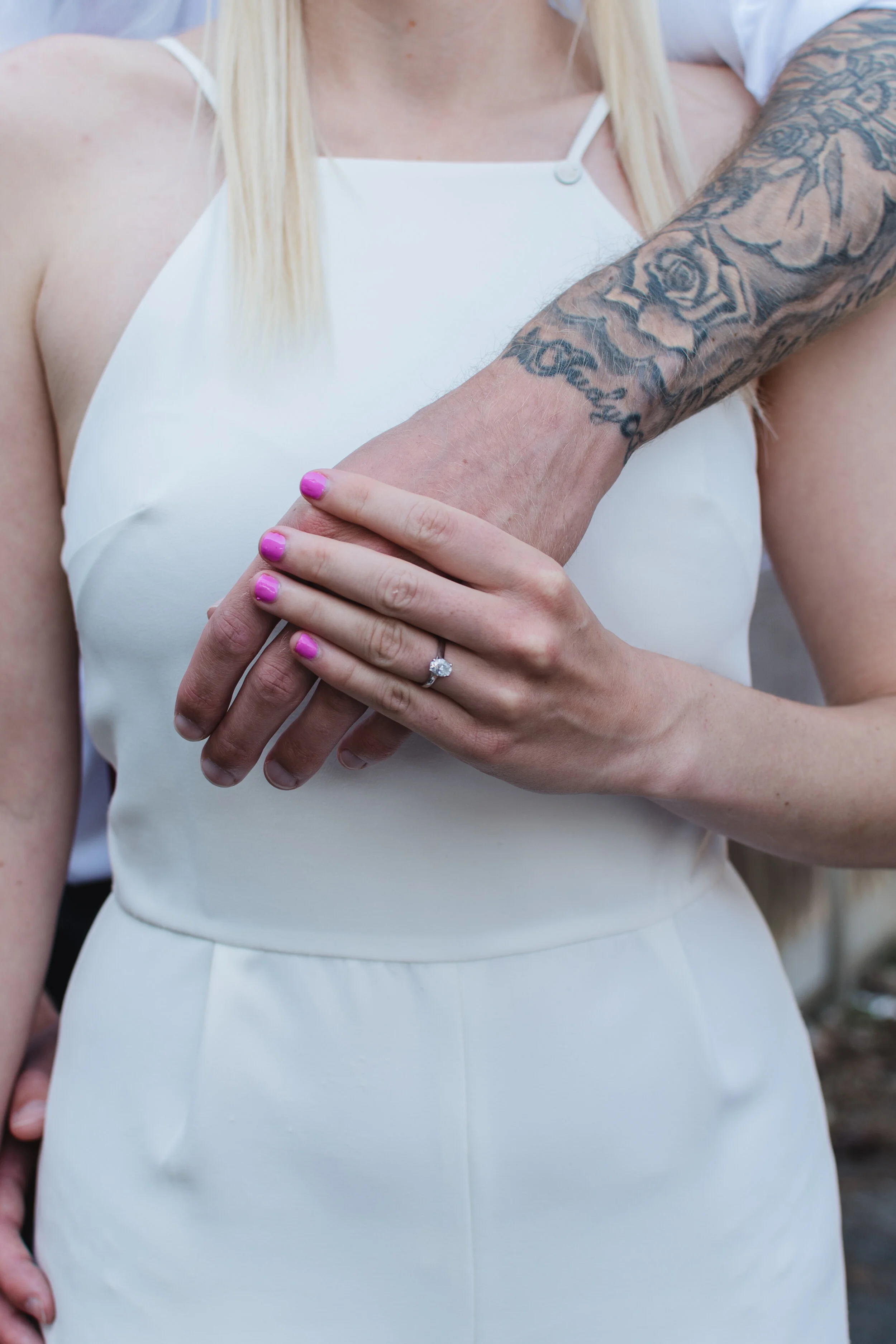 Close-up of two people with arms wrapped, one with a tattooed arm, wearing a wedding ring, and pink nail polish, both in white clothing.