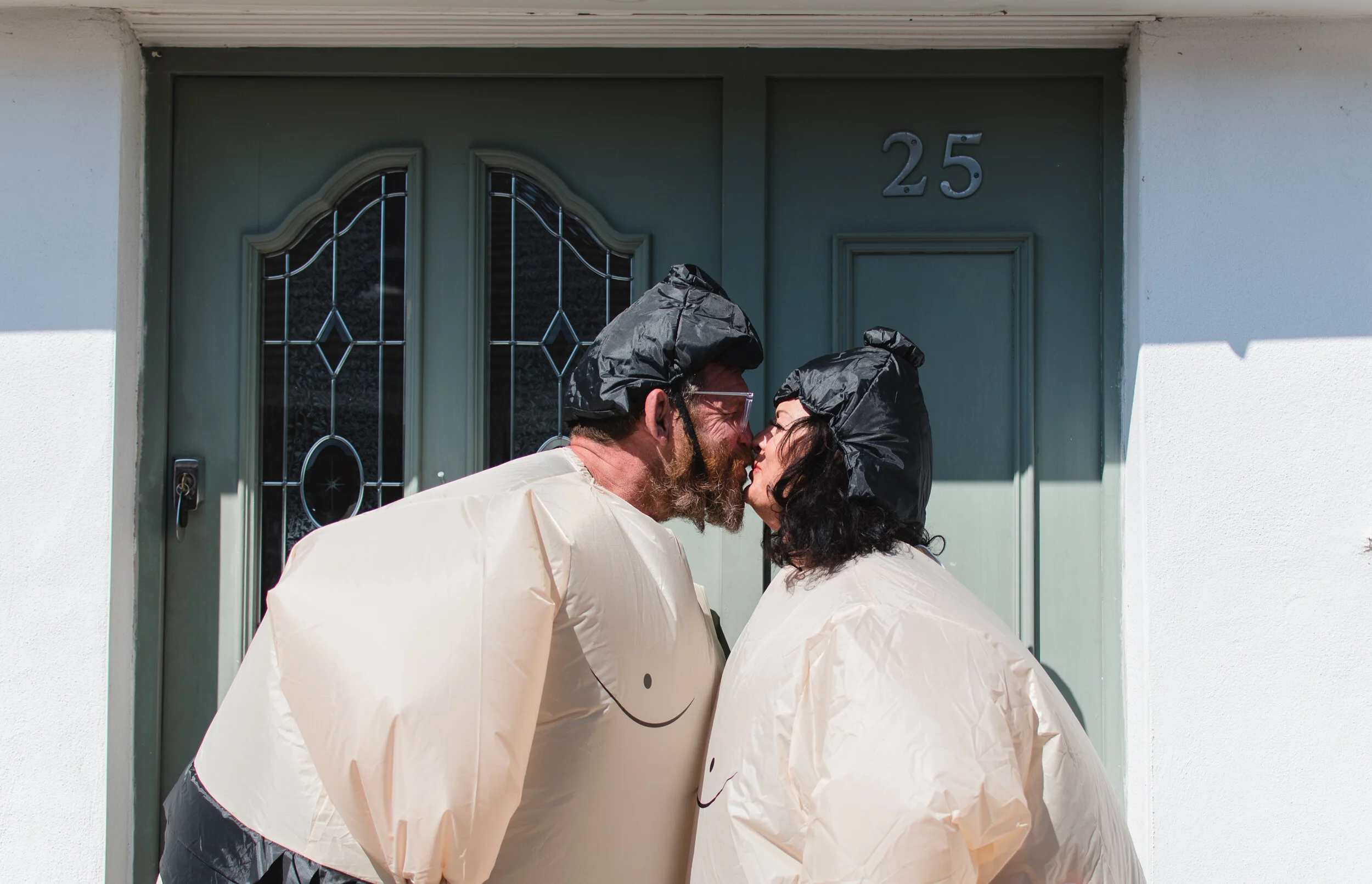 Two people wearing inflatable sumo wrestler suits and hats, standing close to each other in front of a green door with the number 25.