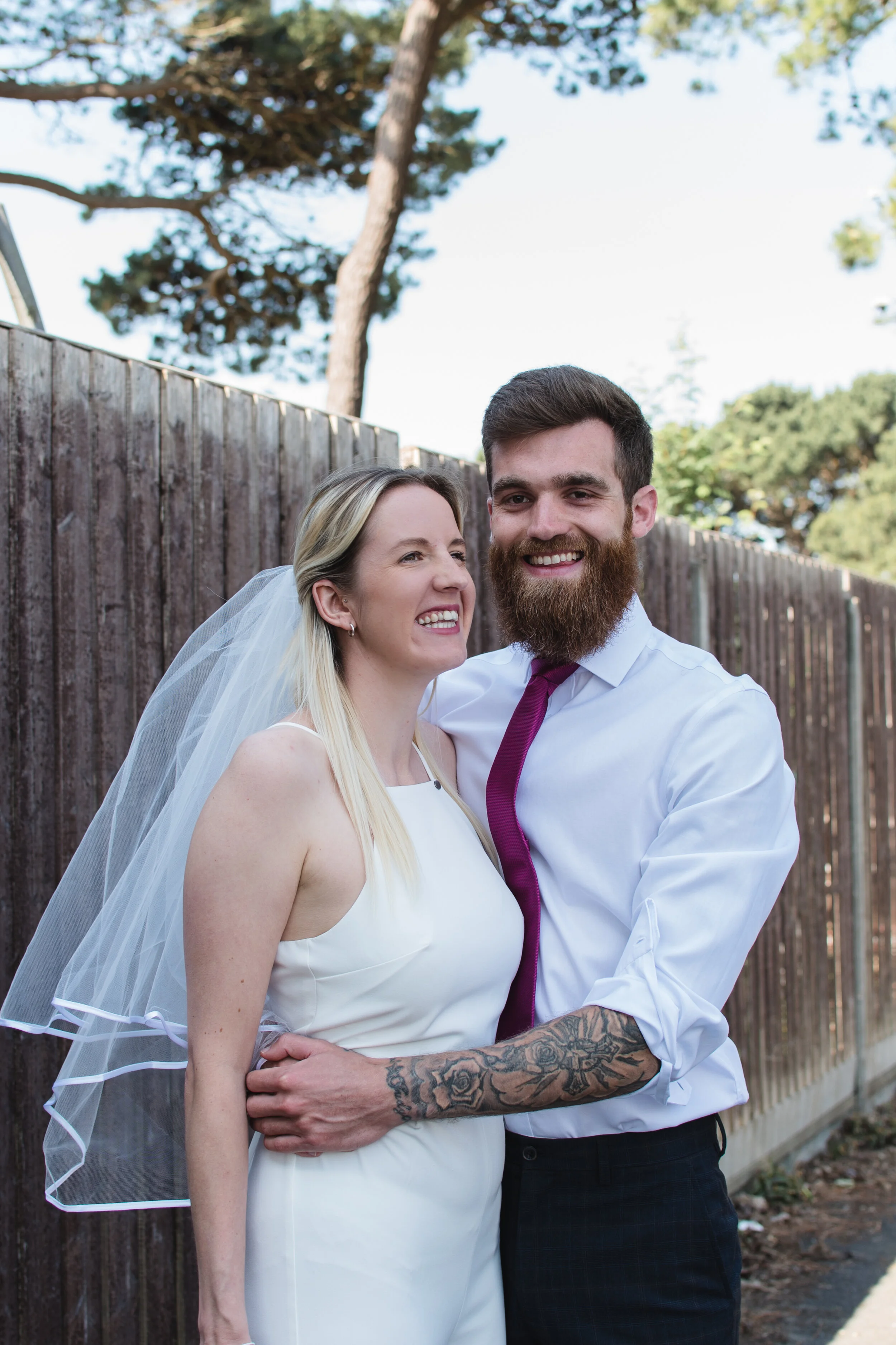 Smiling couple in wedding attire, with the woman wearing a white dress and veil, and the man in a white shirt with a tie, standing outdoors by a wooden fence.