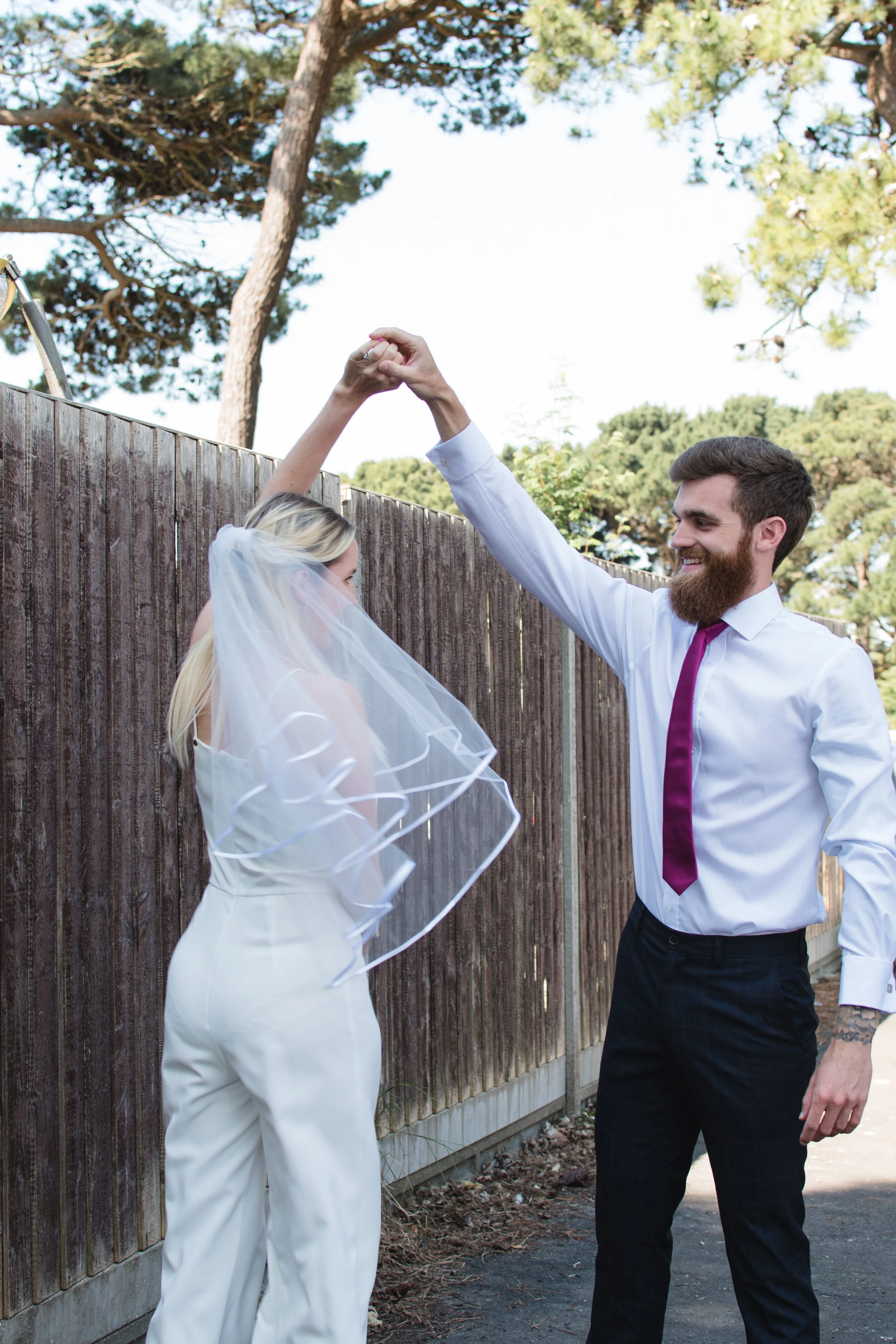 A bride and groom dancing outdoors by a wooden fence, with the bride wearing a white veil and pants and the groom in a white shirt and red tie.