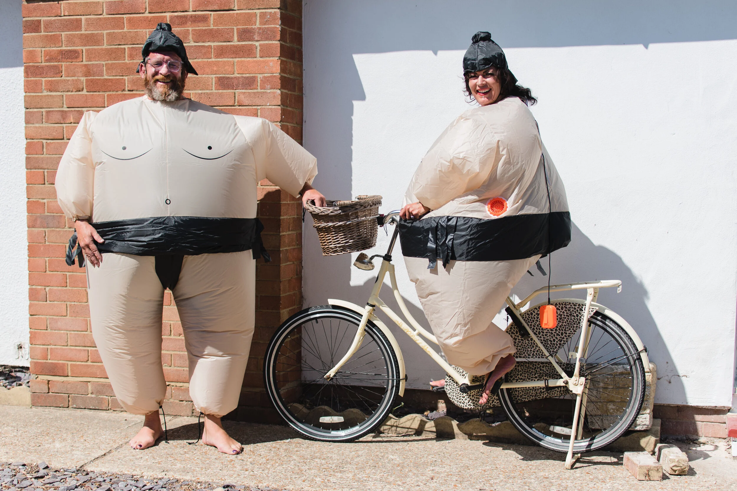 Two people in inflatable sumo wrestler costumes, one standing and the other sitting on a bicycle with a wicker basket, against a brick and white wall background.
