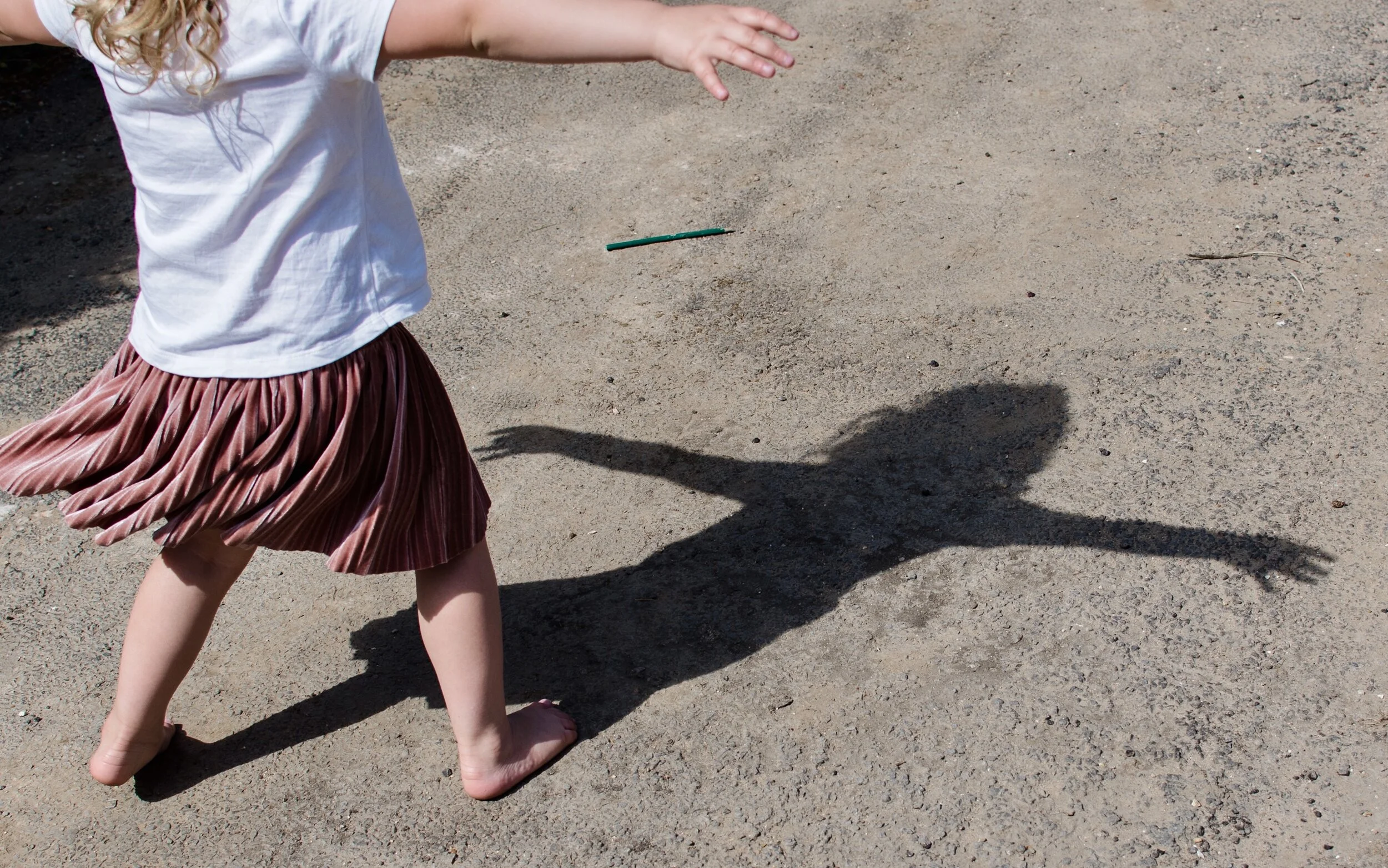 Child in a white shirt and pink skirt dancing on a dirt path, casting a shadow on the ground.