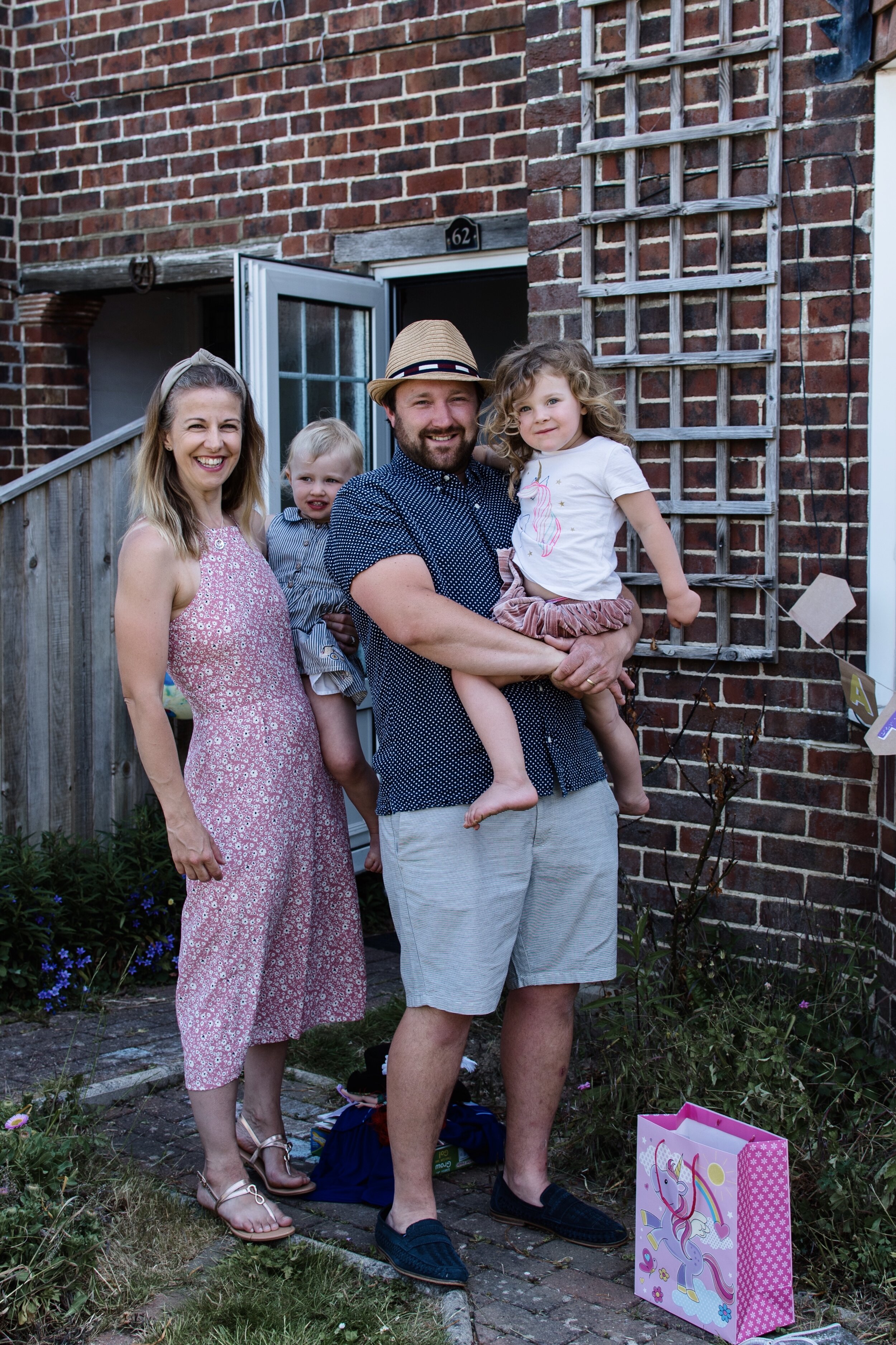 A smiling family standing outside a brick house with an open door. The man is holding a child, and a gift bag with a unicorn design is on the ground beside them.