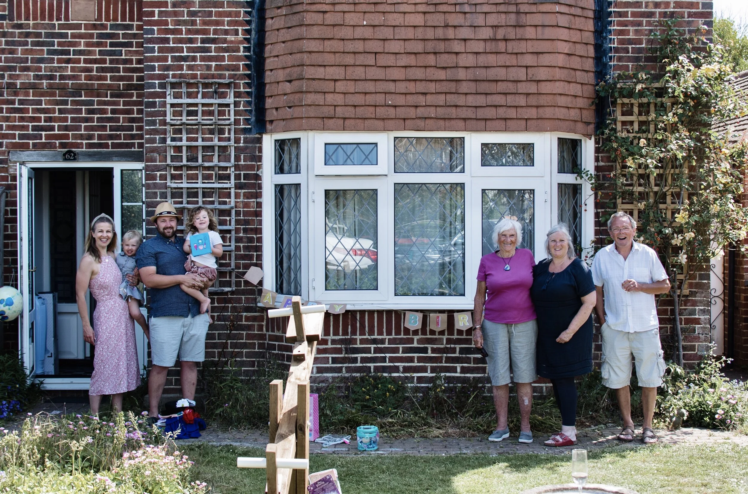 A family group posing in front of a brick house with a bay window, celebrating a birthday with a banner spelling 'HAPPY BIRTHDAY'. There are flowers in the foreground and three adults with two children on the left, and three adults on the right.