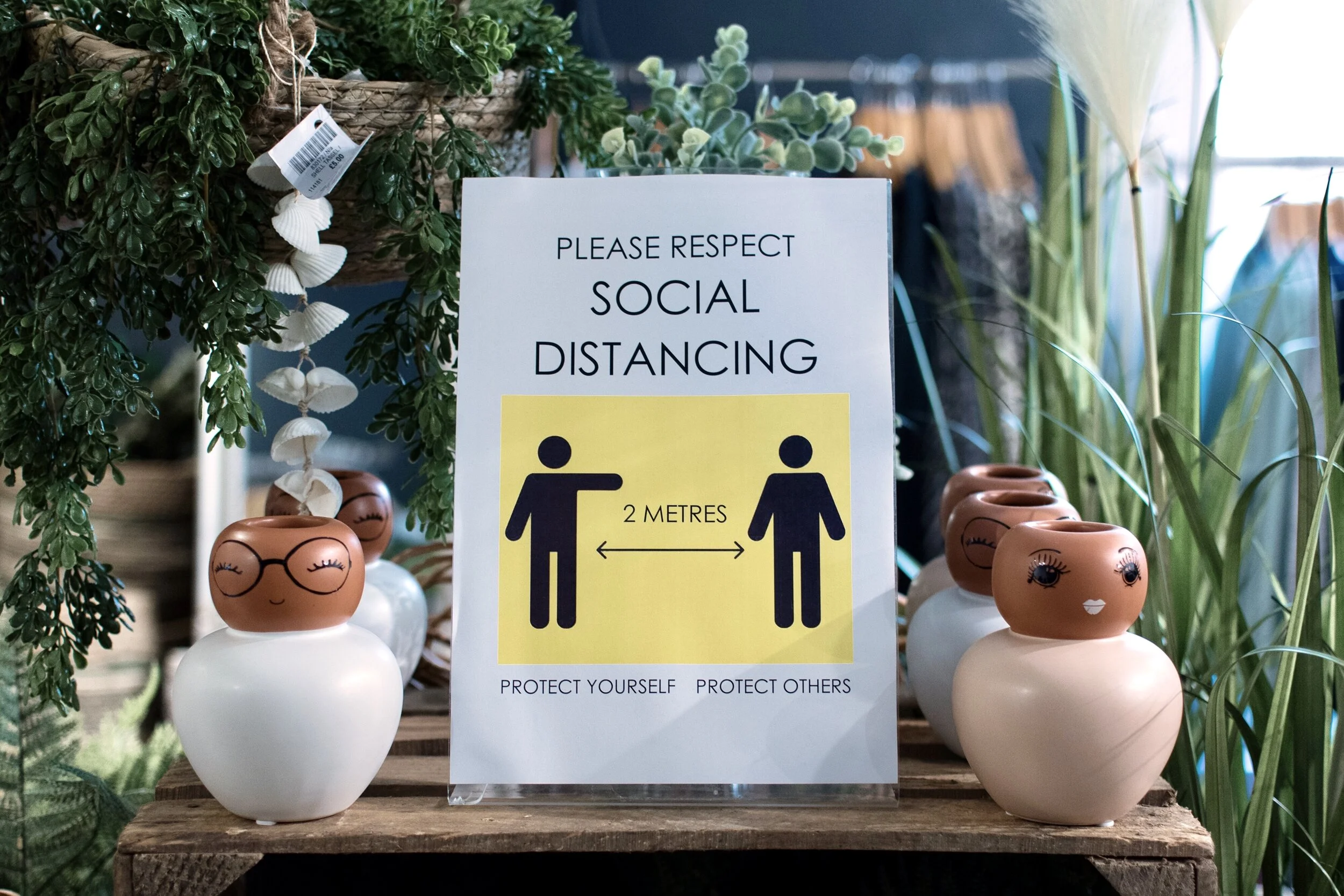 A social distancing sign in a store, surrounded by decorative planters with faces, with greenery in the background.