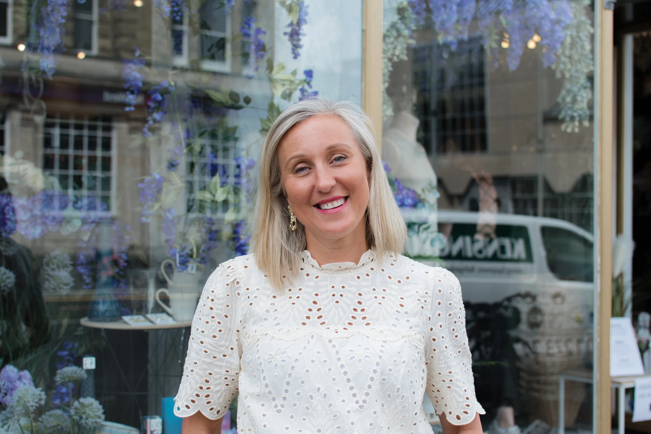 Woman smiling outside a shop with floral window display.