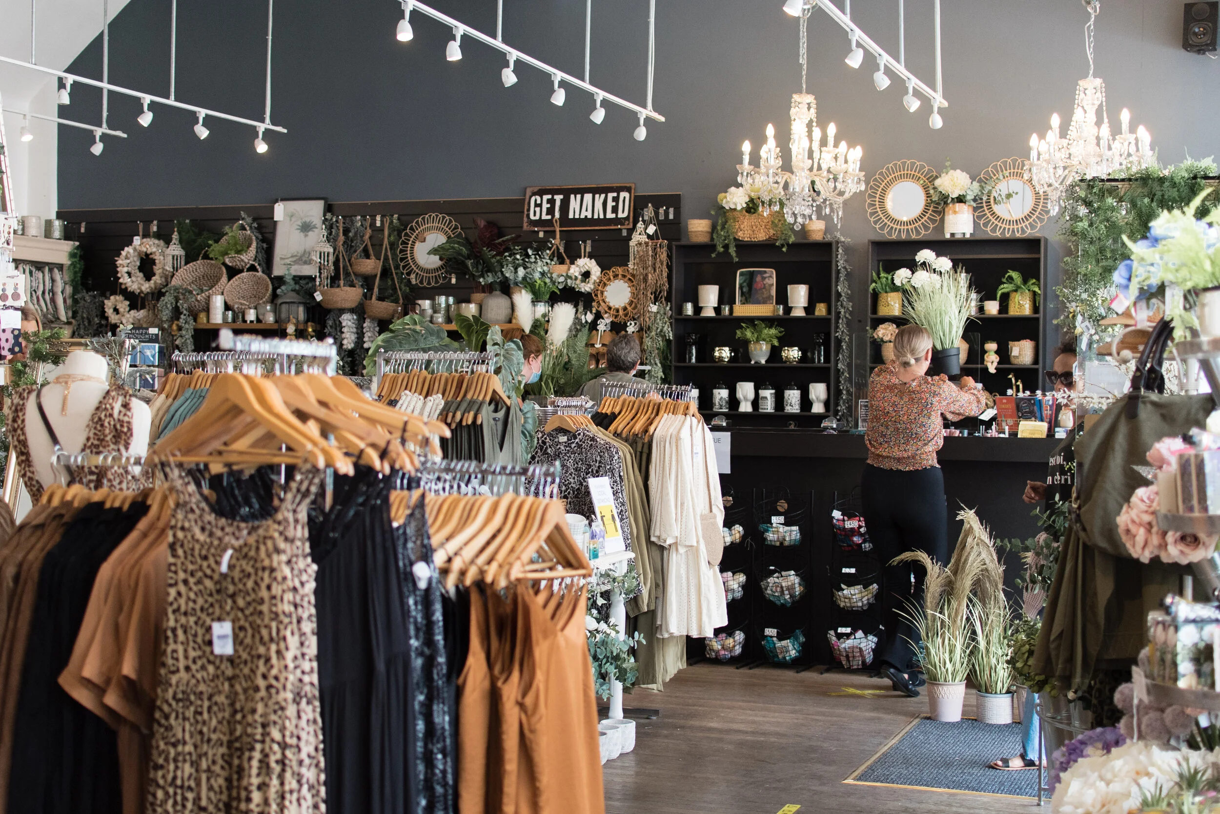Interior of a boutique store with clothing racks and home decor items. A person stands at the checkout counter surrounded by plants, baskets, and a "Get Naked" sign. Chandeliers hang from the ceiling.
